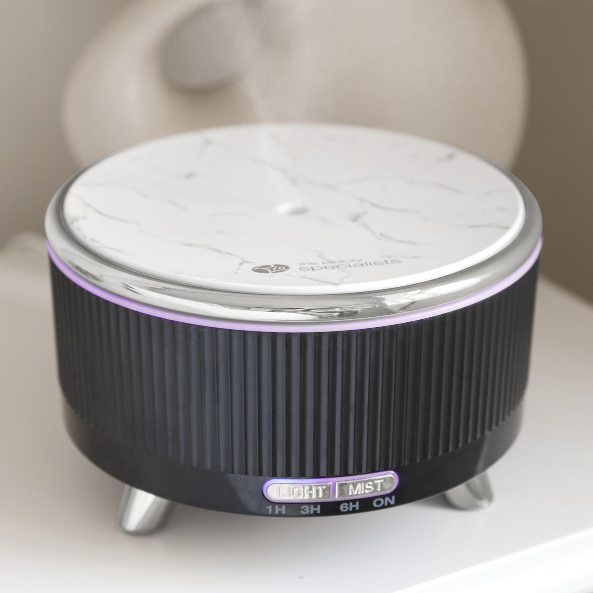 The Rio SONAS Aroma Diffuser, Humidifier & Night-Light features a white marbled top, black ribbed sides, metallic legs, and purple mood lighting. On the front are control buttons for Light, Mist, and timer settings. It rests on a white surface.