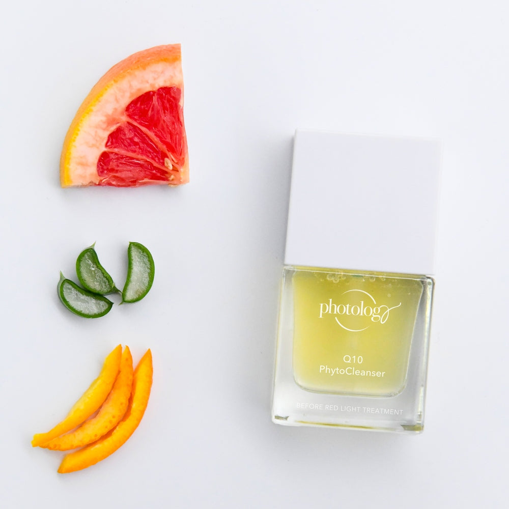 A bottle of Rio Photology Q10 Pre-treatment Phyto Cleanser rests on a white surface, with grapefruit, aloe vera, and mango slices artfully arranged in the top left corner.