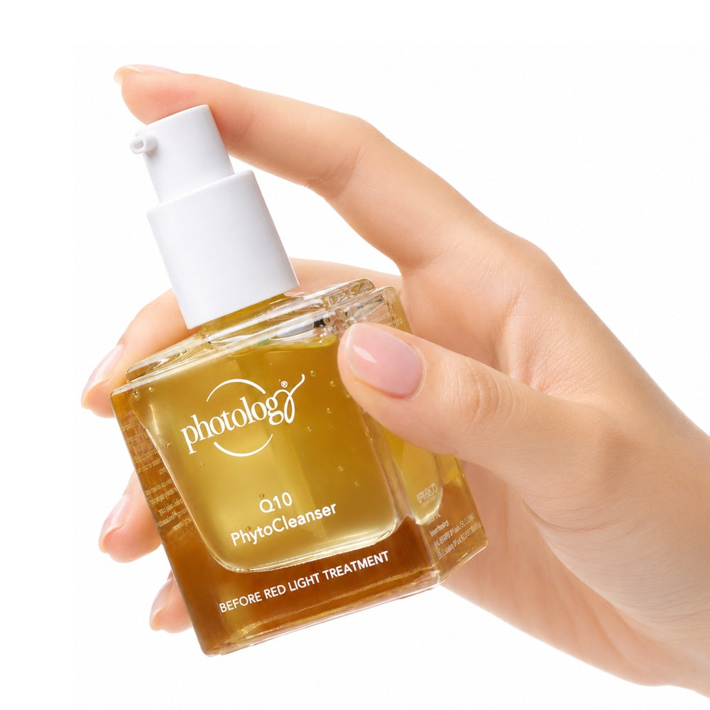 A manicured hand holds a square glass bottle of Rio Photology Q10 Pre-treatment Phyto Cleanser, filled with yellow liquid. The label reads BEFORE RED LIGHT TREATMENT, highlighting its use before red light therapy.