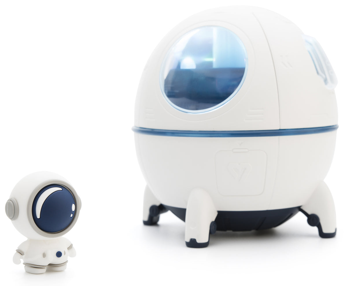 A small toy astronaut stands beside a larger, white Rio Spaceship Explorer Childs Essential Oil Diffuser, Humidifier and Night Light with blue windows and four short legs, both displayed against a white background.
