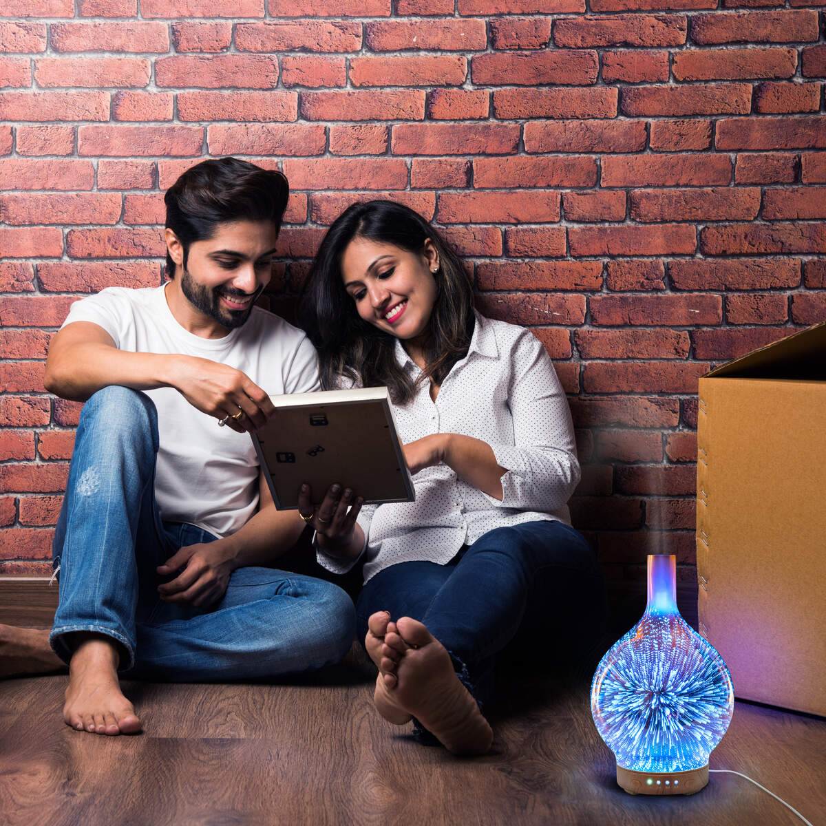 A smiling couple sits on a wooden floor against a brick wall, looking at a tablet. An open box and the Rio ELLA Aroma Diffuser, Humidifier and Night-Light create a relaxing atmosphere with its gentle glow.