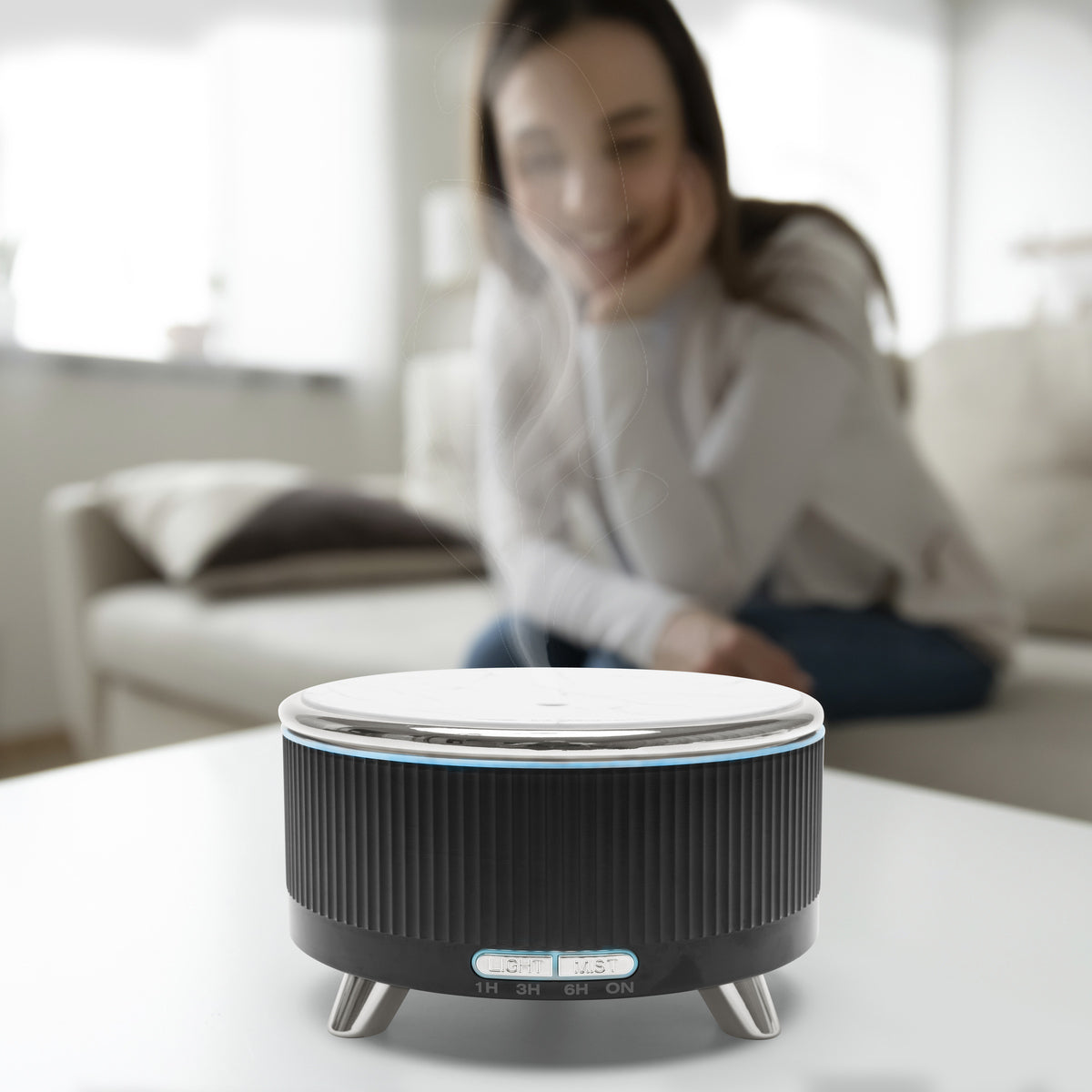 A woman sits on a couch in the background, smiling at a Rio SONAS Aroma Diffuser, Humidifier & Night-Light in sleek black and silver, emitting soothing moisture on the table in the foreground.