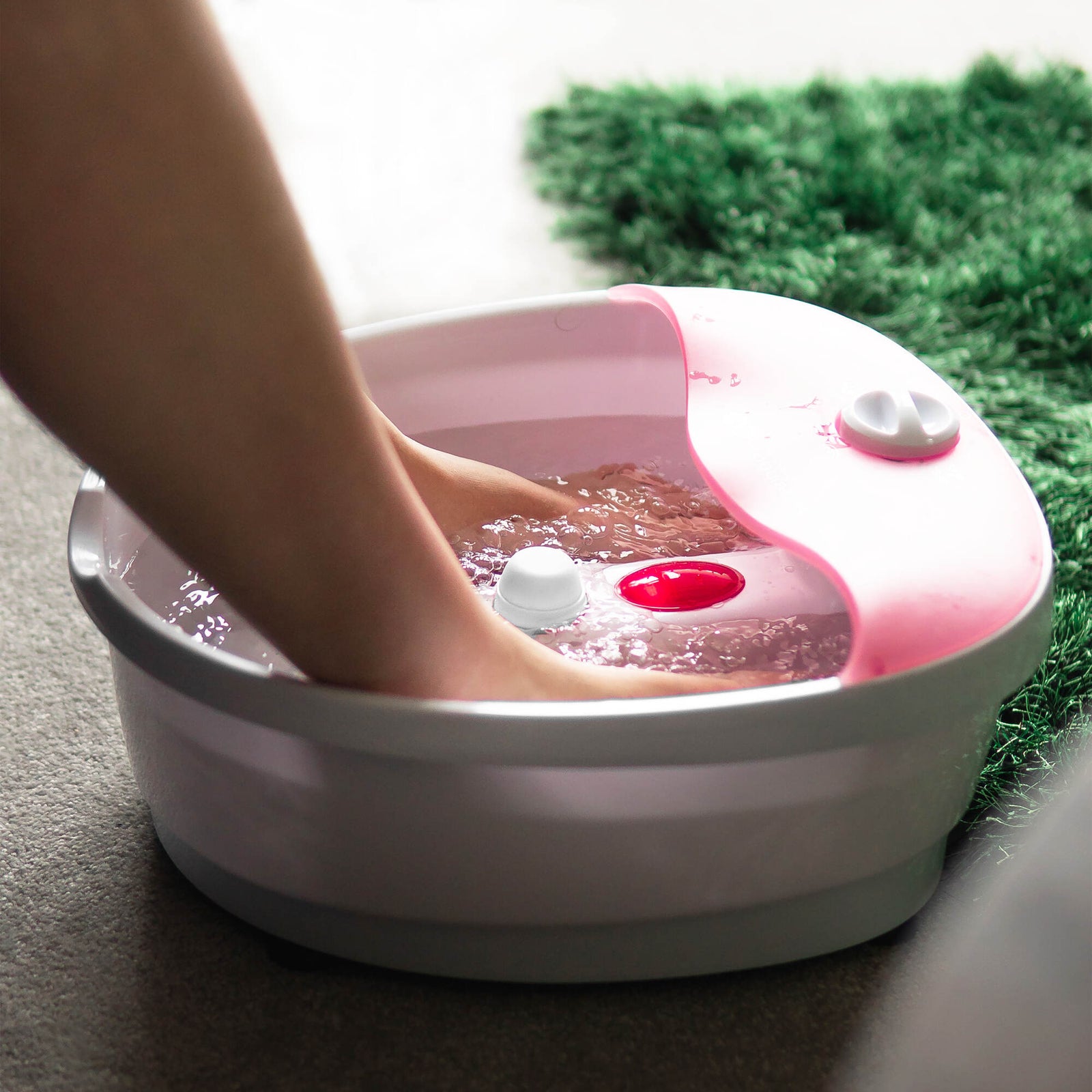 A person enjoys a relaxing soak with the Rio Soothing Waves Foot Bath Spa & Massager, feet immersed in its pink and white basin filled with water, placed on a carpeted floor near a green rug—pure at-home indulgence.