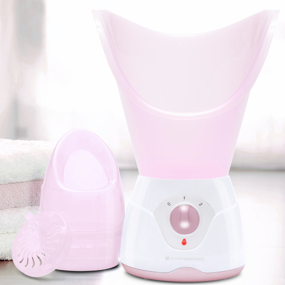 The Rio Facial Sauna Skincare with Steam Inhaler in pink, featuring a dial and detachable parts, is placed near folded towels in a softly lit room.