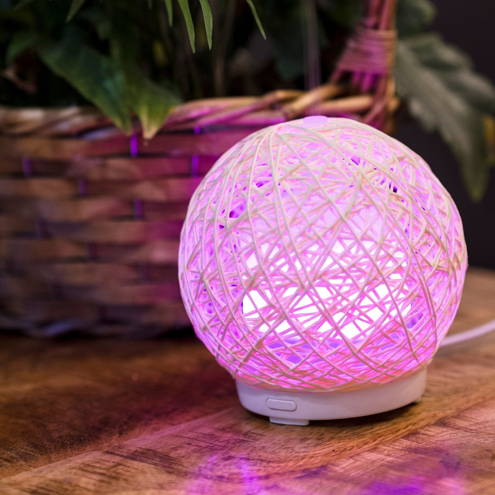 The Rio Lola Aroma Diffuser, Humidifier, and Night Light adds a tranquil glow with its spherical woven design and purple light, perfect for creating a serene atmosphere on your wooden surface beside greenery.