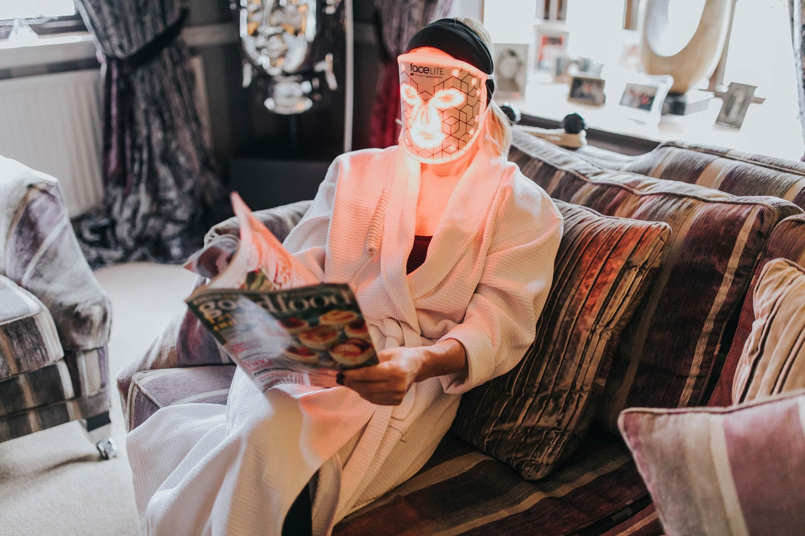 lady relaxing at home on sofa reading magazine wearing facelite beauty boosting LED face mask with red and infrared lights illuminated