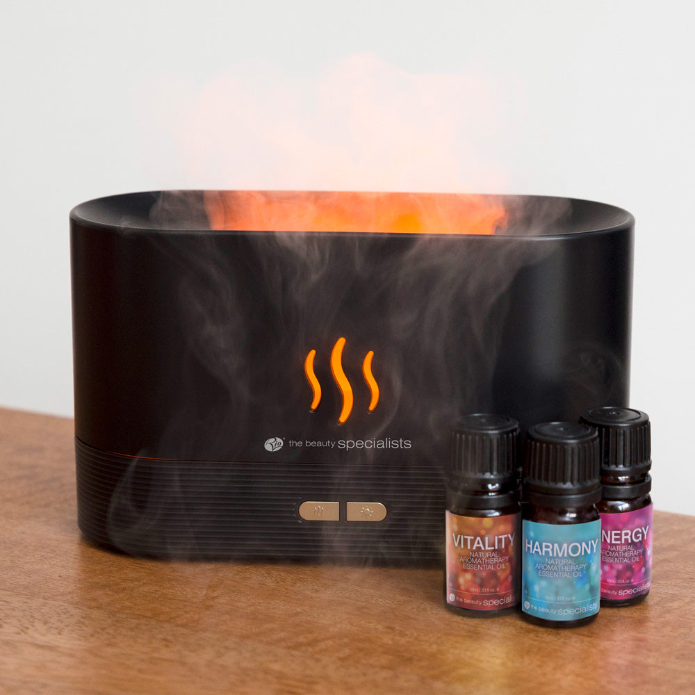 The Rio ALTA Aroma Diffuser, Humidifier, and Night-light in black emits an orange glow and mist, with three essential oils—Vitality, Harmony, and Energy—displayed beside it on a wooden surface.