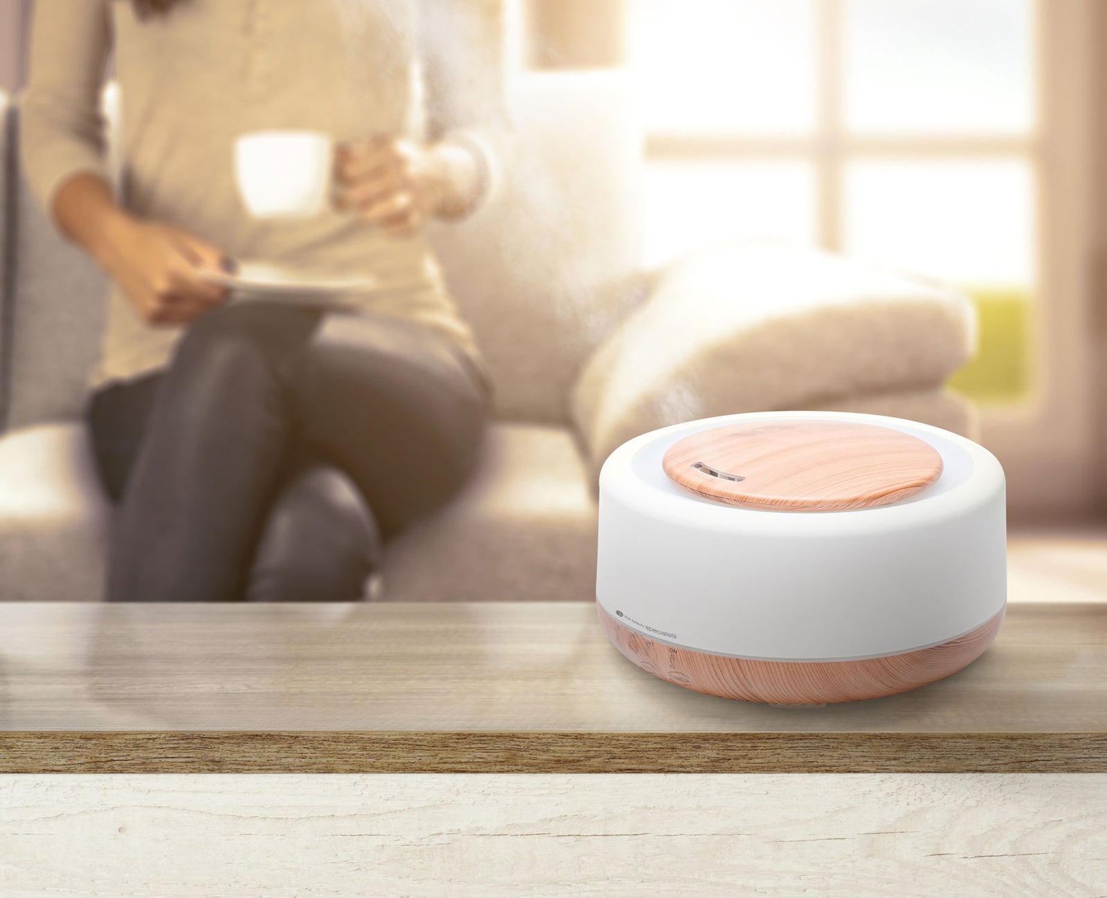 Aroma diffuser placed on a wooden table with female in background drinking a cup of coffee