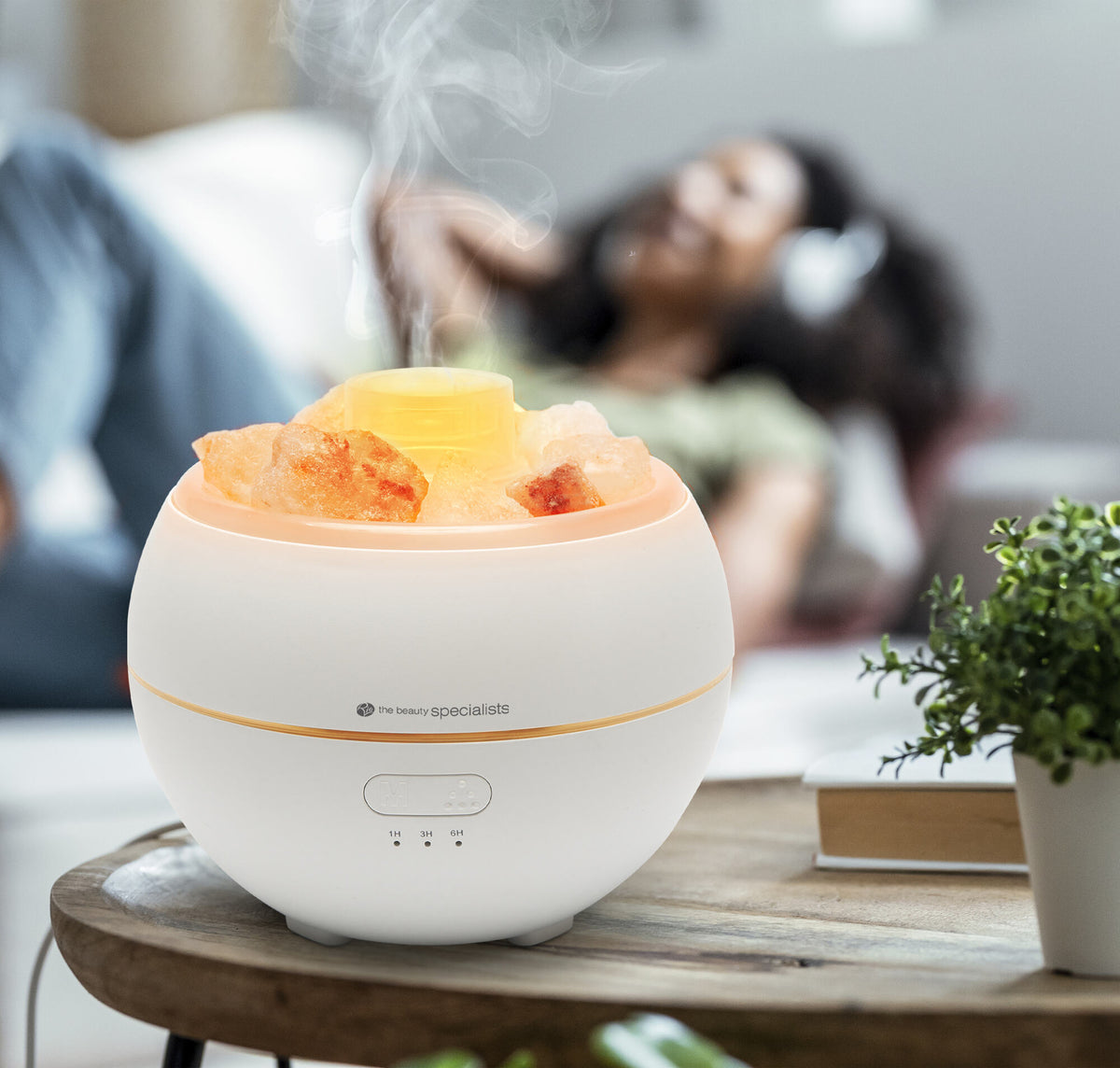 The Rio Aroma Diffuser, Humidifier & Night-Light with Himalayan Rock Salt emits steam on a table, while someone relaxes in the background with headphones.