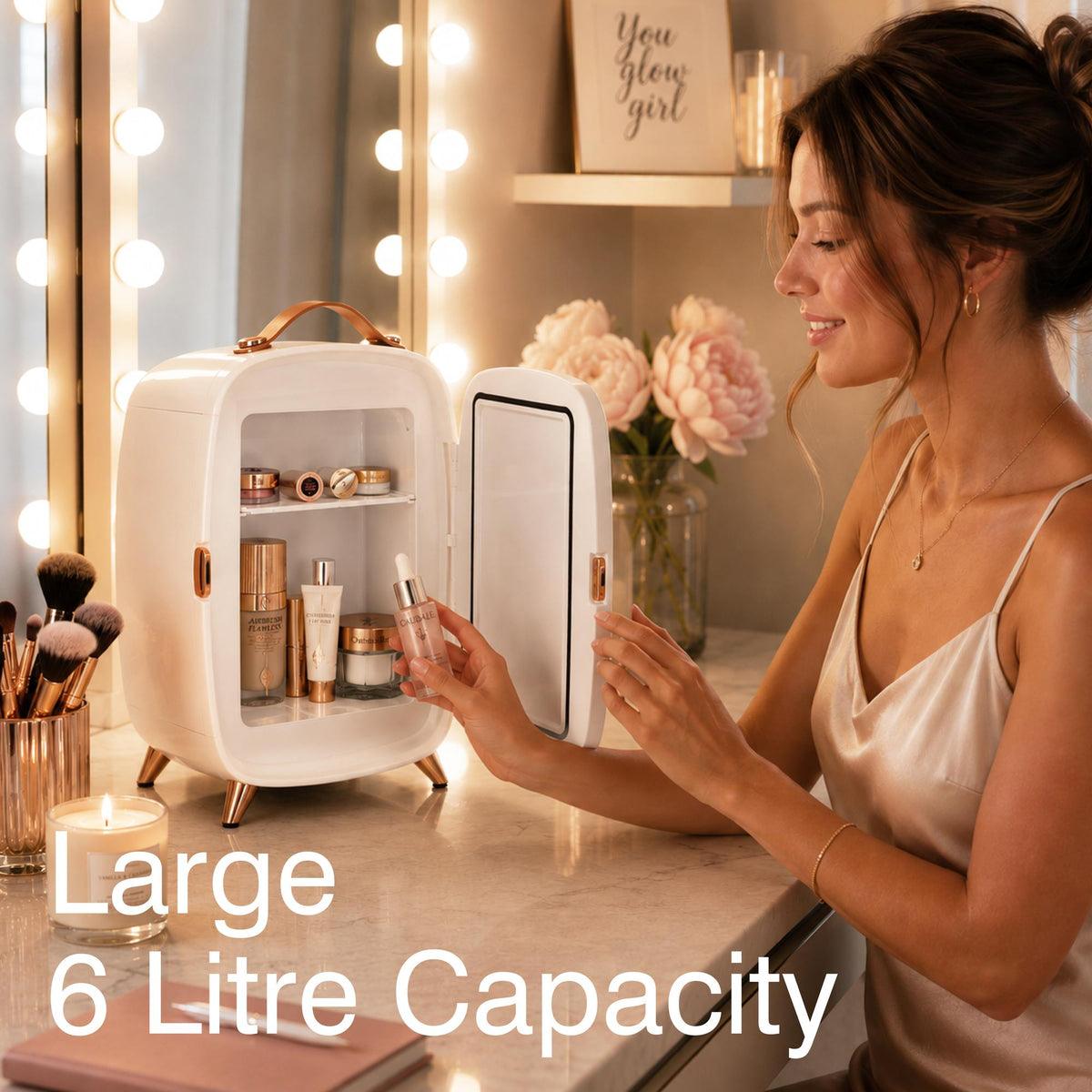 A woman in a satin dress sits at a vanity table adorned with cosmetics and flowers, placing a skincare product into the Rio Beauty Skincare Fridge With Mirror—featuring a large 6-litre capacity and illuminated by lights around the mirror—ideal for keeping creams and other skincare essentials perfectly chilled.