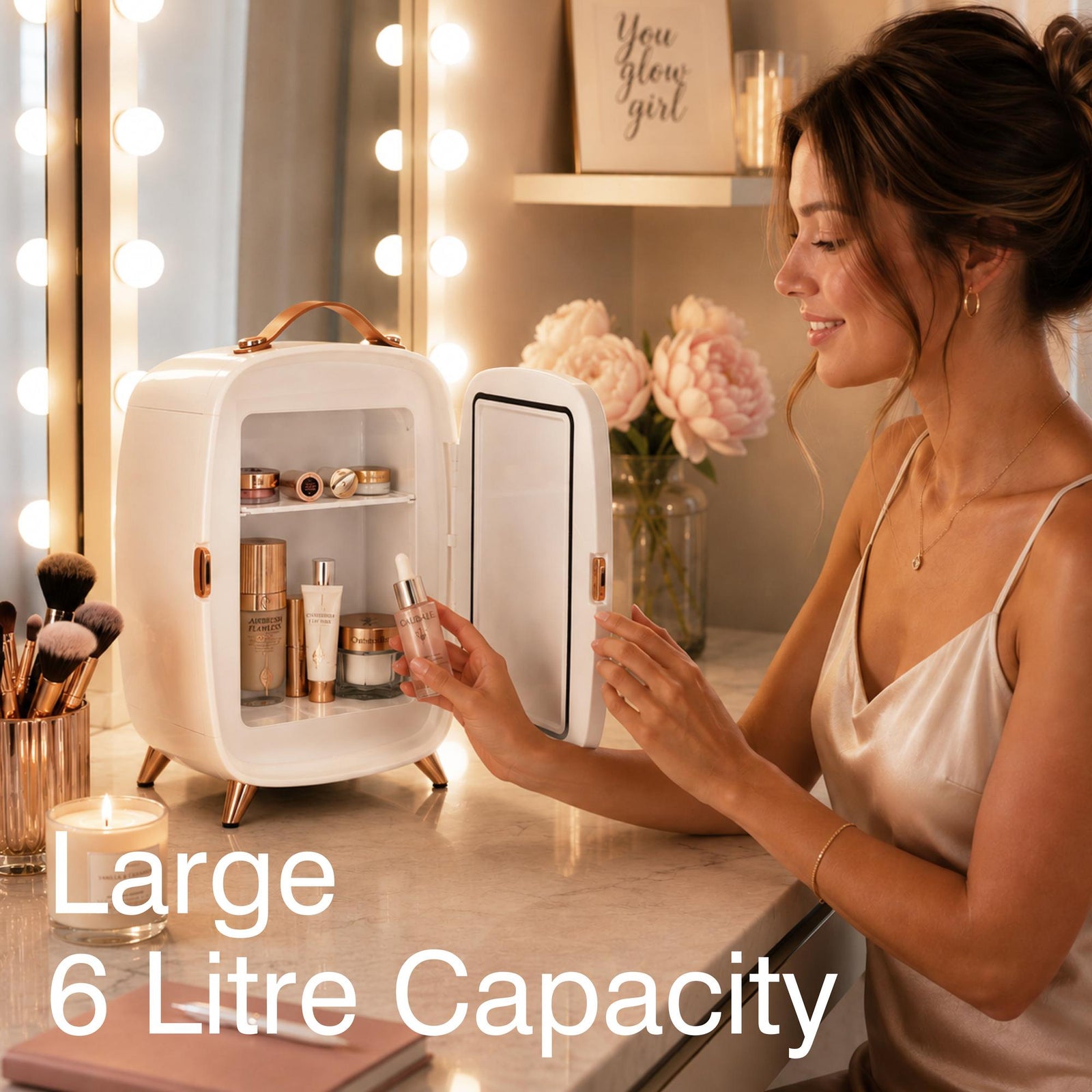 A woman in a satin dress sits at a vanity table adorned with cosmetics and flowers, placing a skincare product into the Rio Beauty Skincare Fridge With Mirror—featuring a large 6-litre capacity and illuminated by lights around the mirror—ideal for keeping creams and other skincare essentials perfectly chilled.