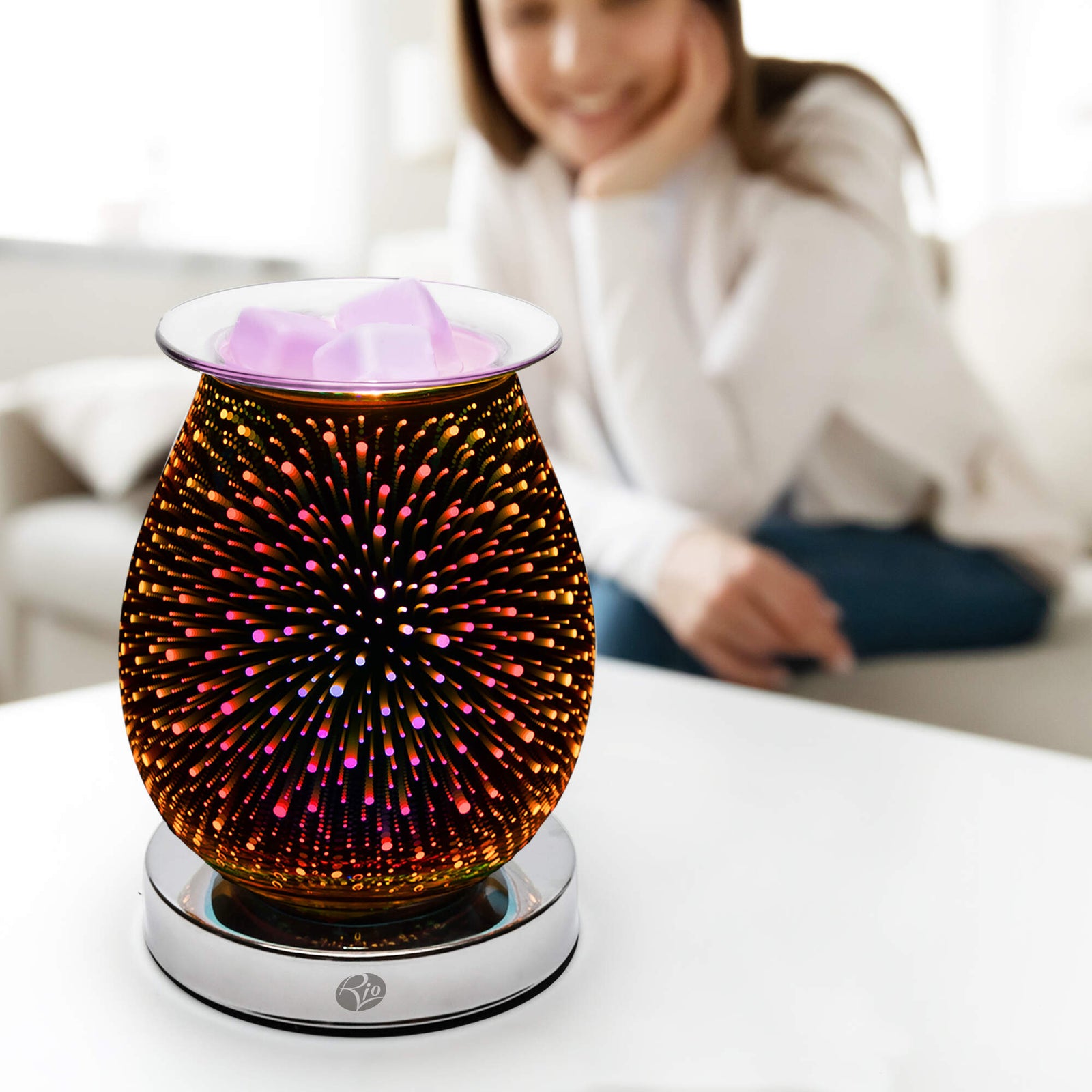 A close-up of the Rio Alura Wax Melt Burner & Aroma Diffuser Lamp, featuring a vibrant firework pattern and glowing warmly on a table. In the background, a woman sits on a couch, smiling gently.