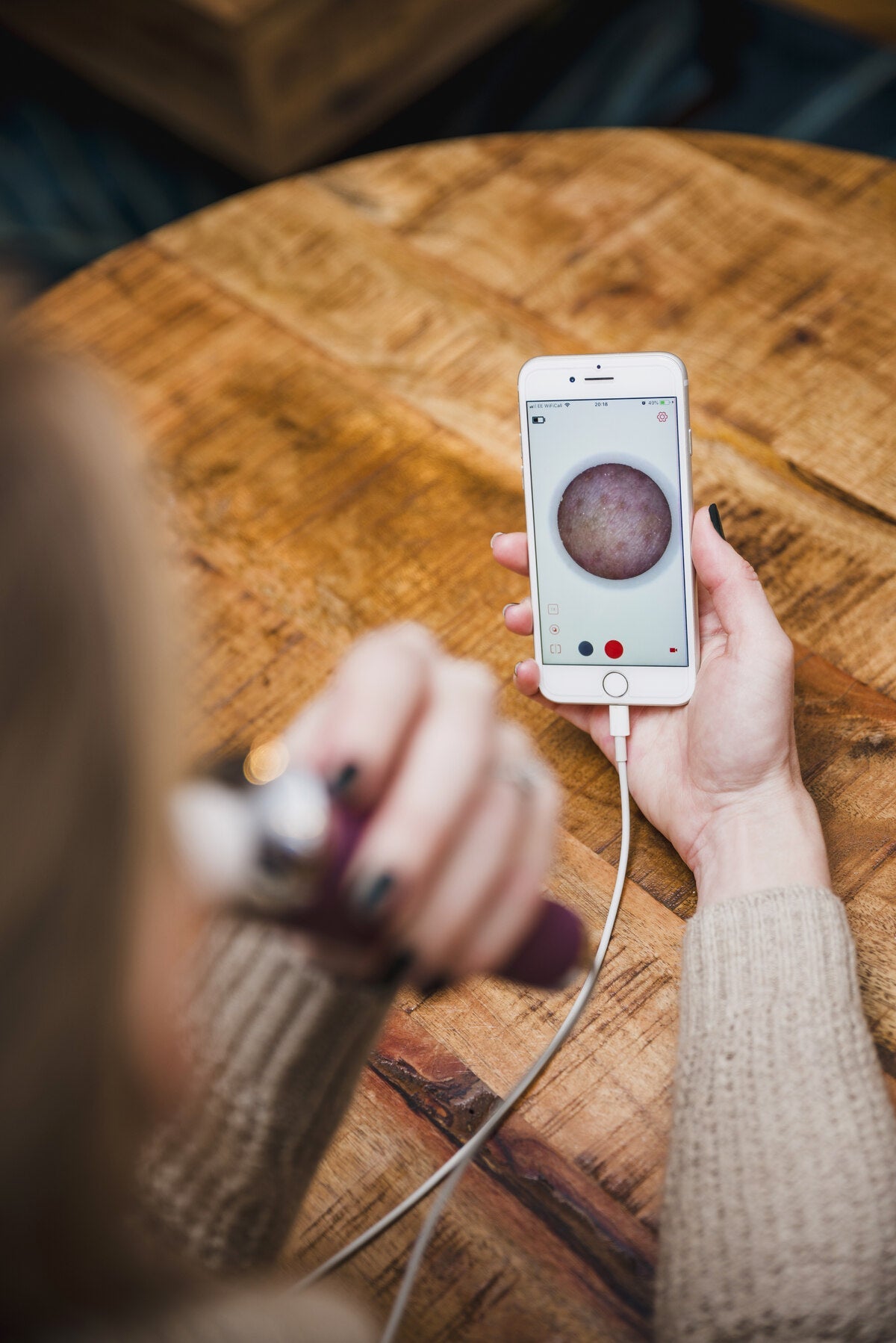 A person uses Rio’s Pore Perfection device with their smartphone to analyze skin up close for personalized skincare, all over a wooden table.