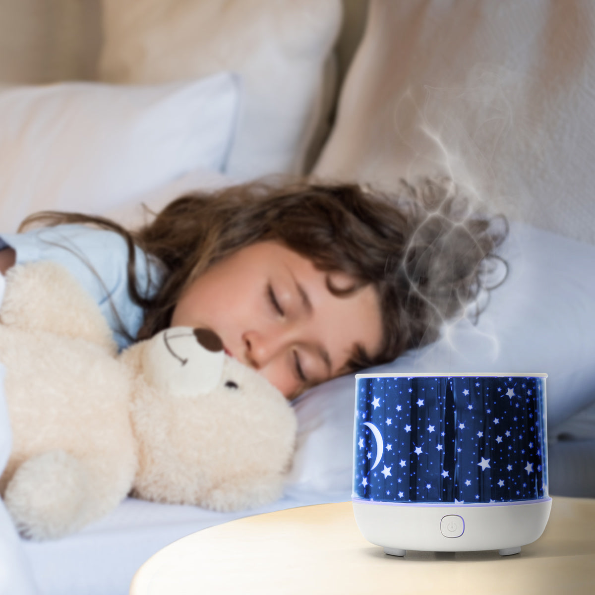 A child cuddles a teddy bear in bed while the Rio Dream Time Aroma Diffuser, Humidifier and Night Light emits soothing mist and gentle light from the nightstand.
