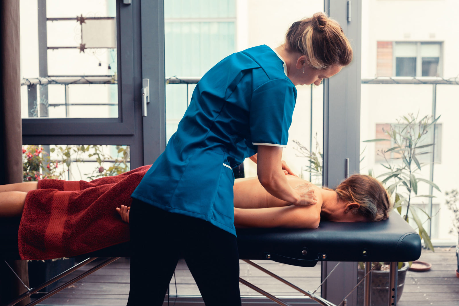 A massage therapist in blue scrubs gives a back massage to a client lying face down, covered with a red towel, on the Rio Professional Aluminium Massage Table & Treatment Couch in a bright room with large windows for an inviting at-home feel.