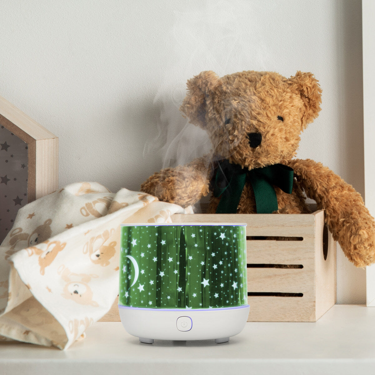 A teddy bear sits in a wooden crate next to a bear-print blanket. In front, the Rio Dream Time Aroma Diffuser, Humidifier and Night Light with star and moon patterns releases visible mist, adding a cozy ambiance.
