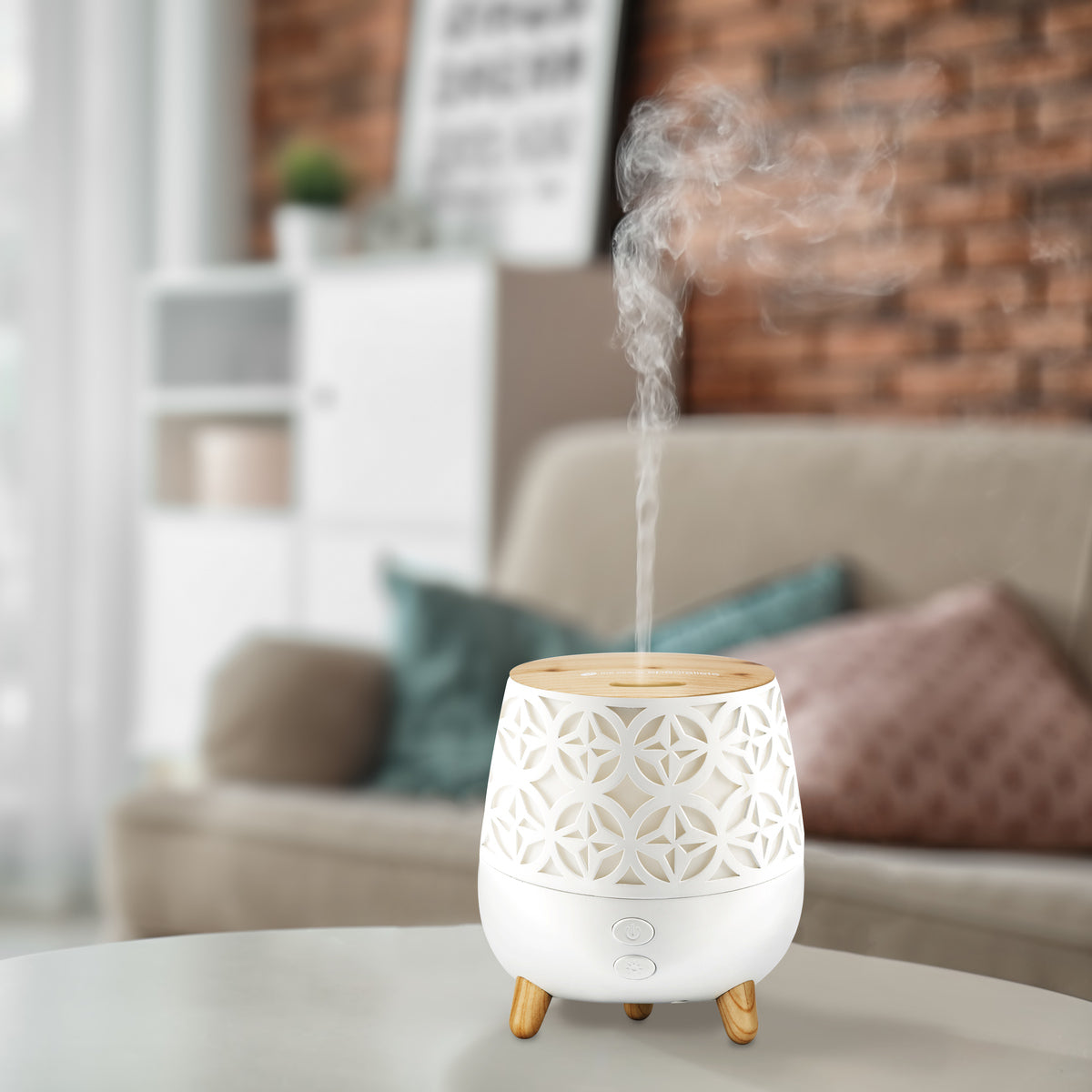 The Rio TALIA Aroma Diffuser, Humidifier & Night-Light in white and wood is shown emitting mist on a round table in a cozy living room with a beige sofa, cushions, and a brick accent wall.