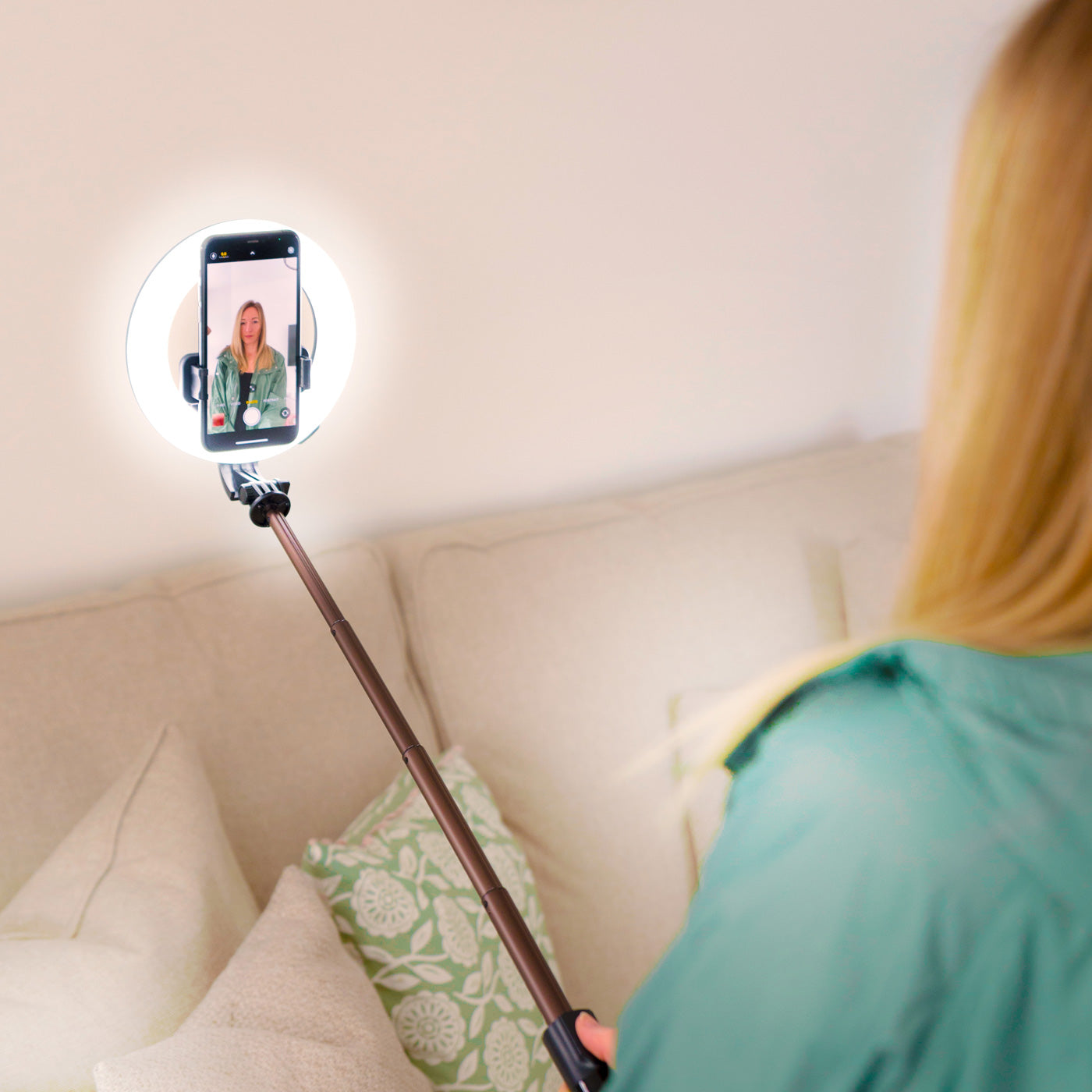 A person with long blonde hair uses the Rio GlamourGlow Pro Beauty & Vlogging Ring Light with a smartphone and selfie stick, vlogging or taking photos while sitting on a beige couch with patterned cushions.