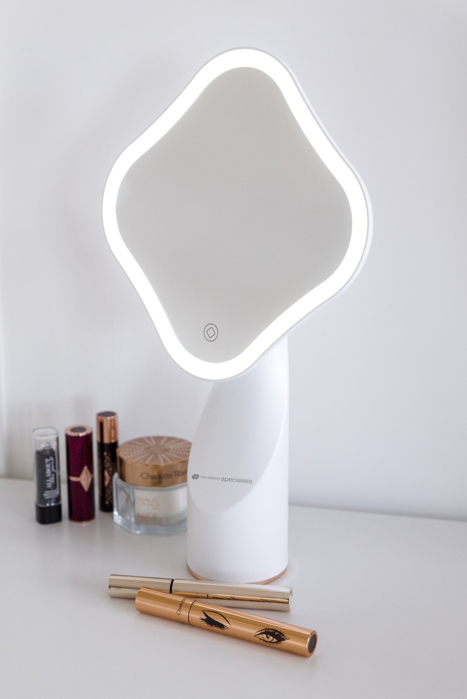 The Rio Illuminated Full Size Beauty Mirror with built-in LED lights sits on a white surface, surrounded by cosmetic items like lipsticks, a jar, and gold mascara tubes. The background is plain and uncluttered.