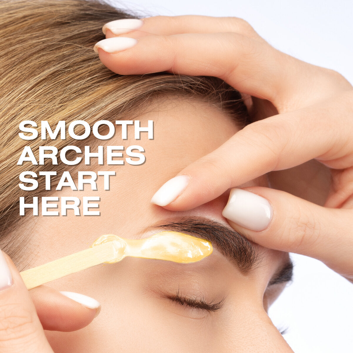 A close-up of a person’s closed eye as another applies Rio Honey Go Smooth Stripless Hard Wax Beads (100g) with a wooden stick to shape their eyebrow. White text reads, “SMOOTH ARCHES START HERE.”.