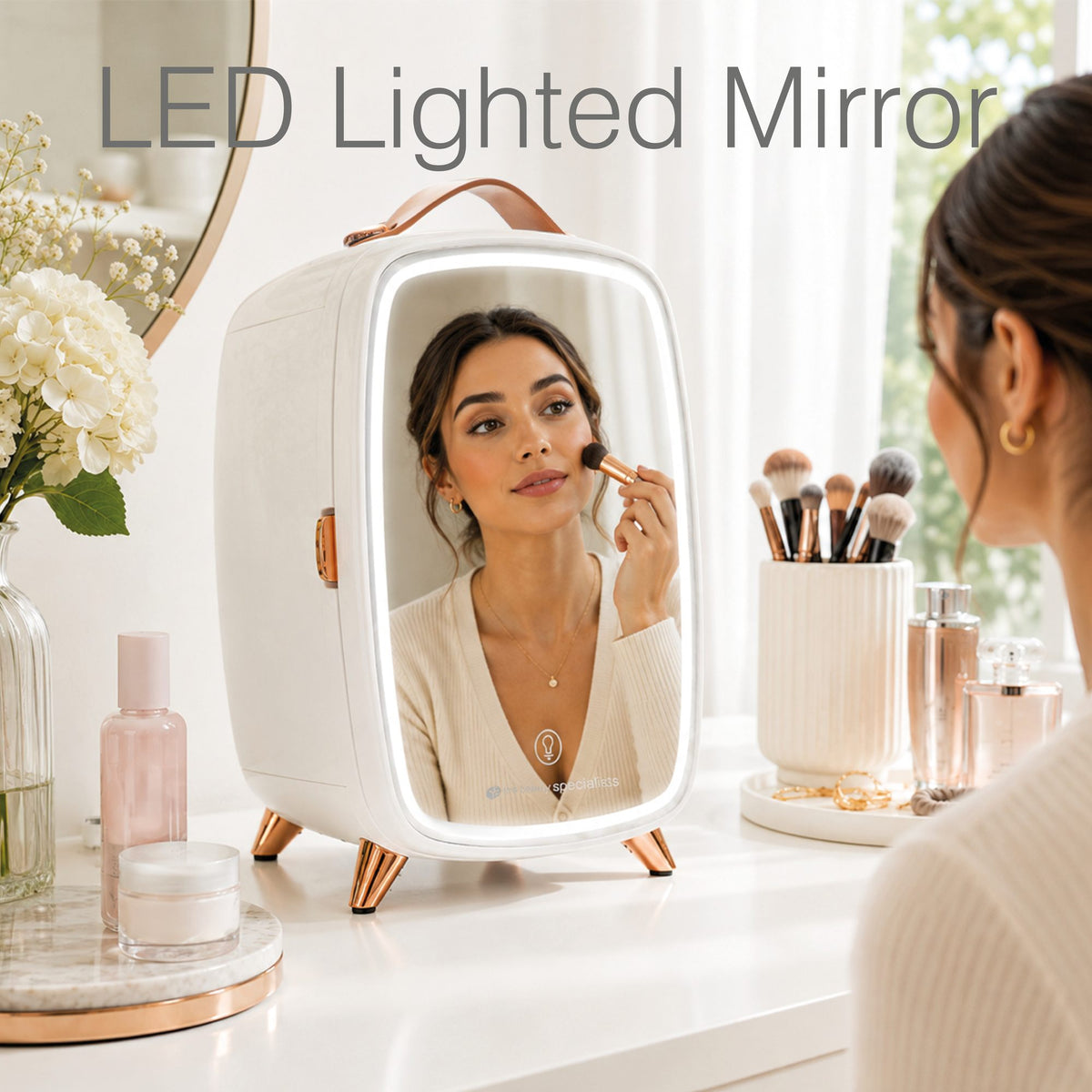 A woman applies lipstick while looking into the mirror on her Rio Beauty Skincare Fridge. Makeup brushes, cosmetics, creams, and flowers are arranged neatly nearby. The skincare fridge features a mirrored door for convenient touch-ups.
