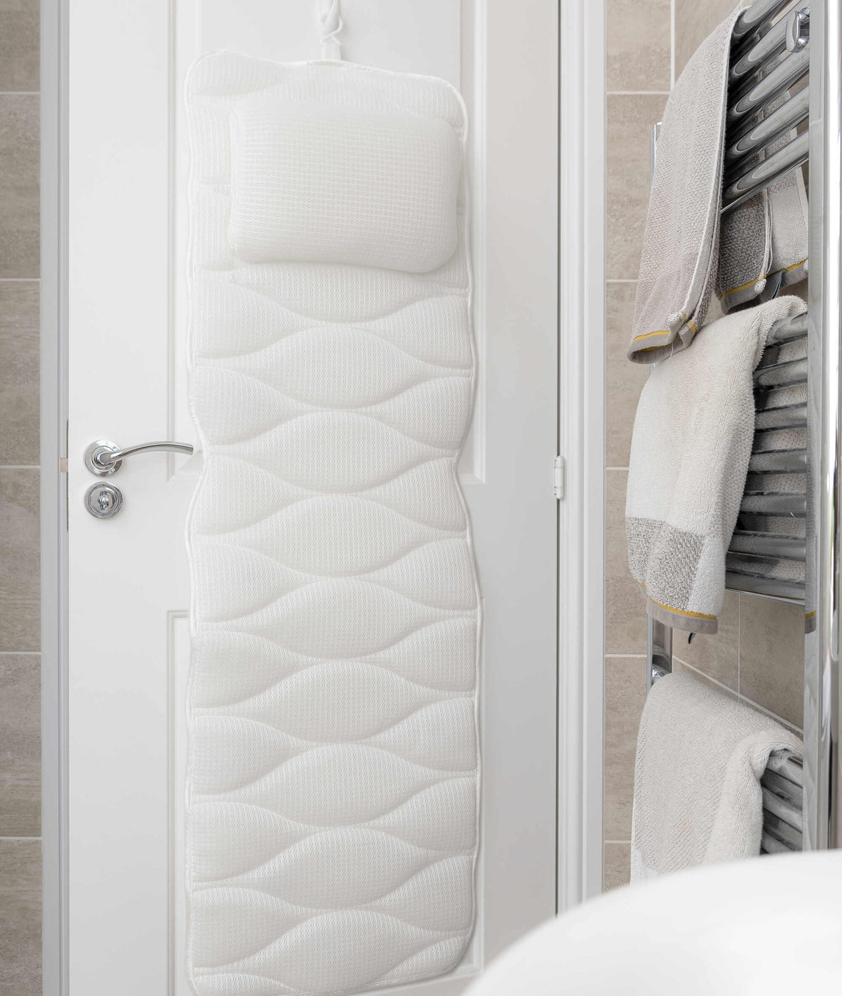 The Rio Luxury Bath Mat & Pillow, featuring a white padded design with headrest, hangs by the door near a heated towel rail with folded beige and white towels, creating an inviting space for bathing relaxation.