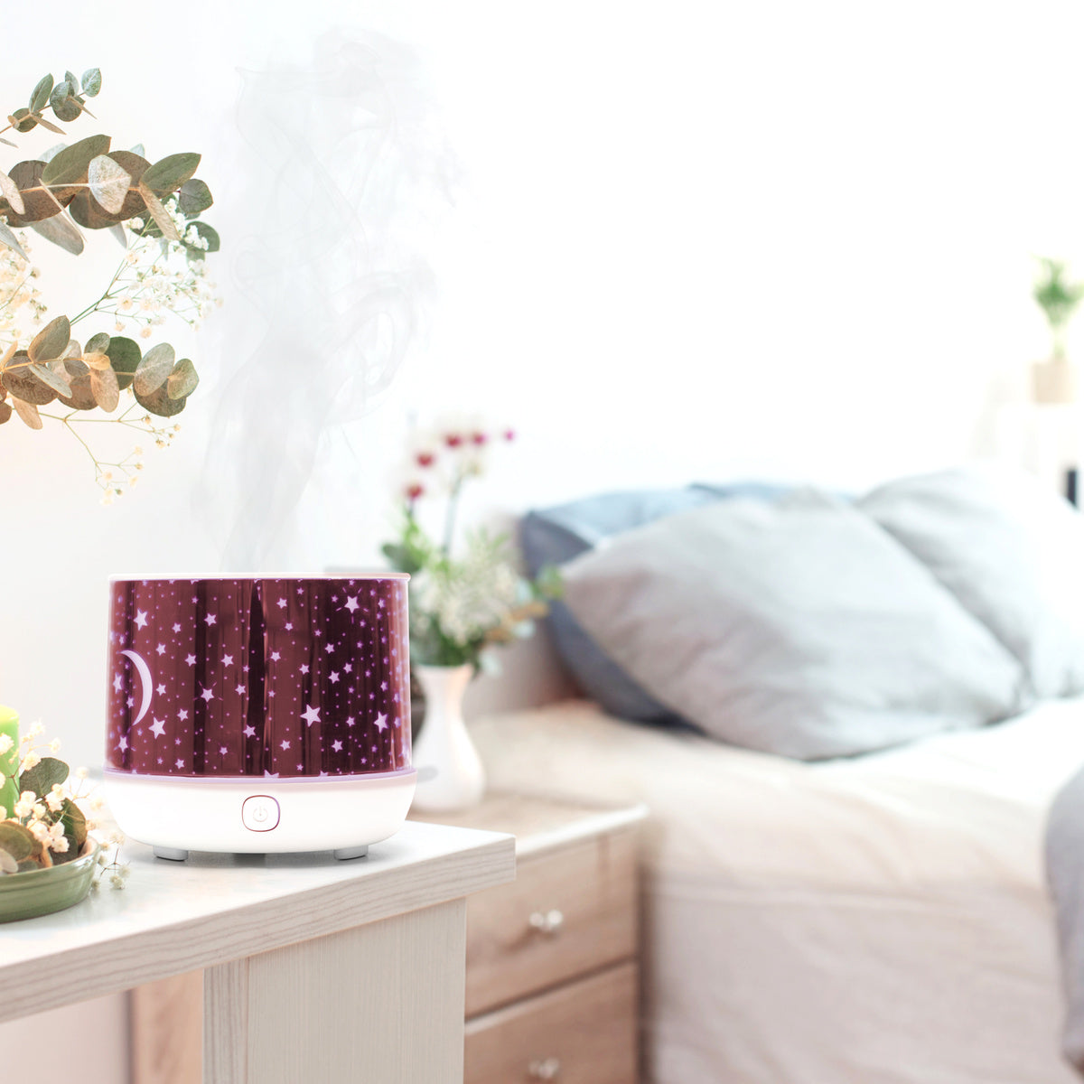 The Rio Dream Time Aroma Diffuser, Humidifier and Night Light with a purple star design and visible mist rests on a bedside table in a cozy, plant-filled bedroom with a neatly made bed in the background.