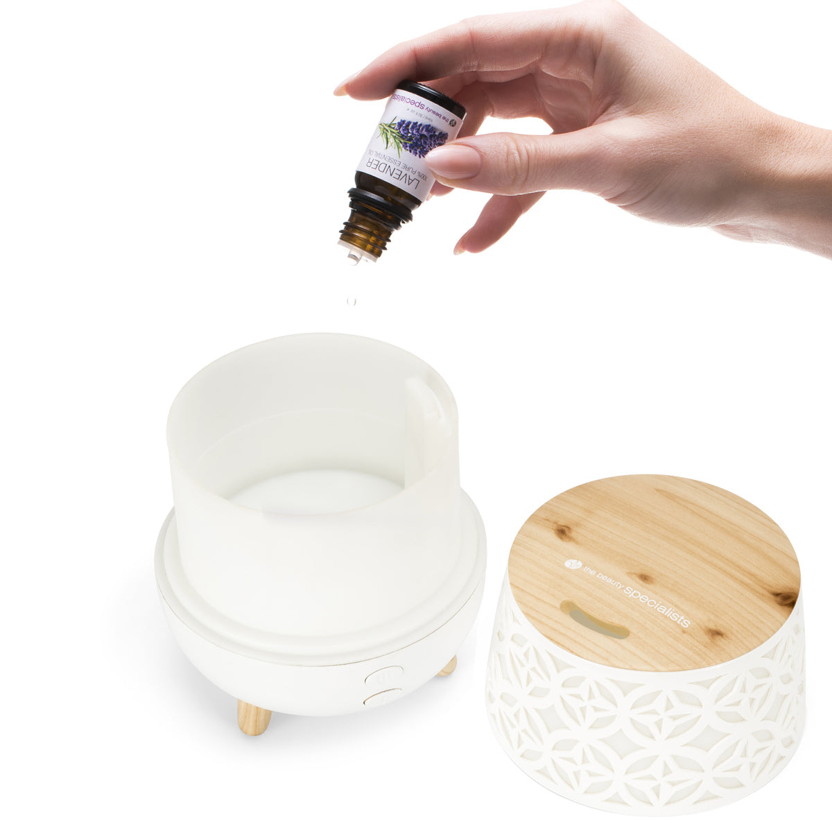 A hand adds drops of lavender essential oil to the TALIA Aroma Diffuser, Humidifier & Night-Light by Rio, shown with its removable wooden lid beside it on a white background.