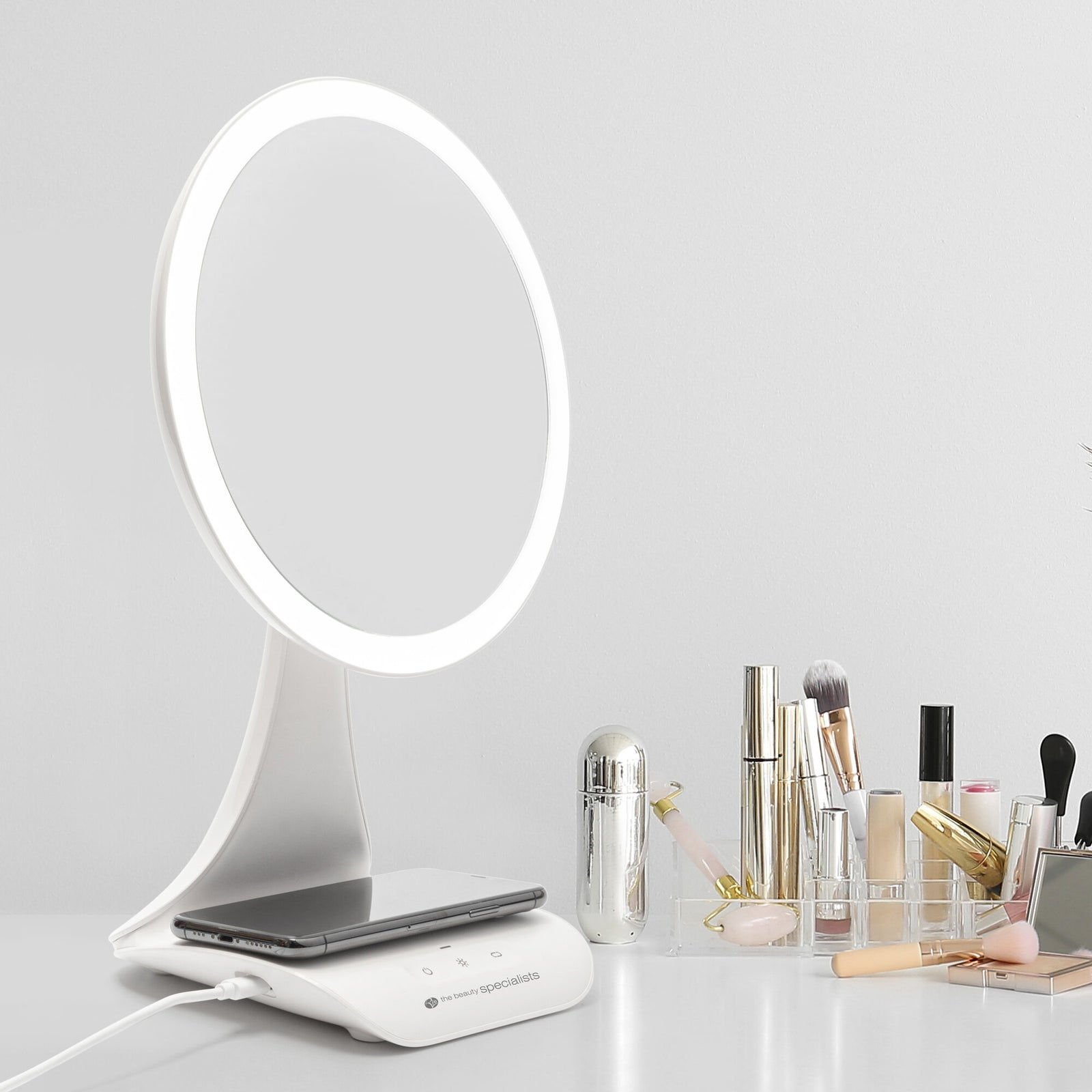 The Rio 5x magnification LED makeup mirror with a wireless charging station sits on a white surface, next to a clear organizer holding brushes and beauty products, against a plain white wall.