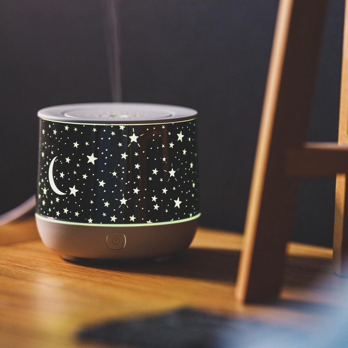 The Rio Dream Time Aroma Diffuser, Humidifier and Night Light features a glowing star and crescent moon design. It emits mist while doubling as a night light, and sits on a wooden surface with part of another wooden object visible nearby.