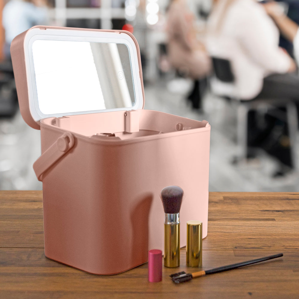 A Rio Lush Box Vanity Case - Large in pink, with its lid open and mirror visible, rests on a wooden table. Makeup items including a brush, lipstick, and mascara are arranged in front, while people are blurred in the background.