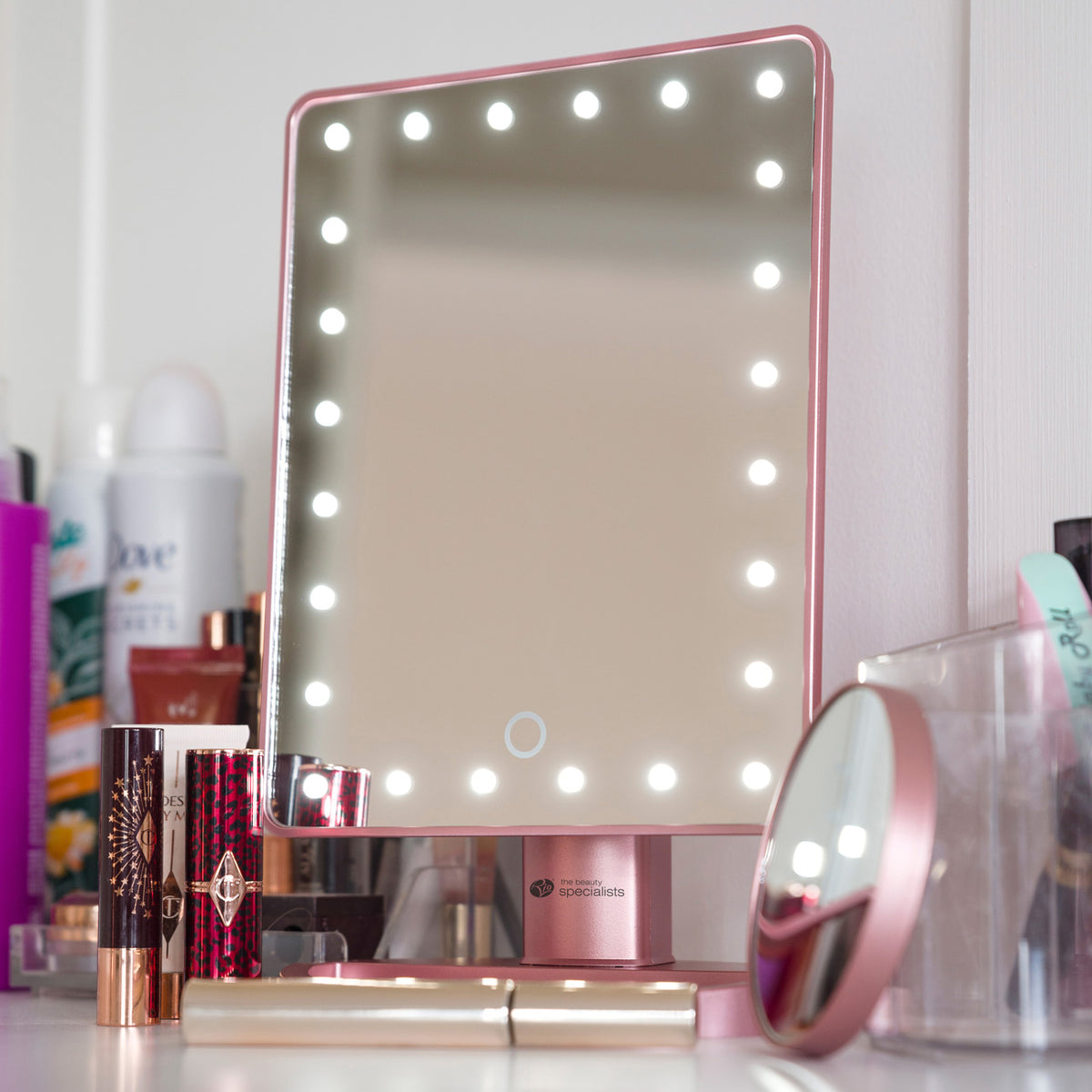 The Rio Rose Gold 24 LED Makeup Mirror with a mini removable 10X Magnifying Mirror sits on a makeup table with lipsticks, foundations, and a compact. In the mirror’s reflection, the magnifying mirror and blurred wall can be seen in the background.