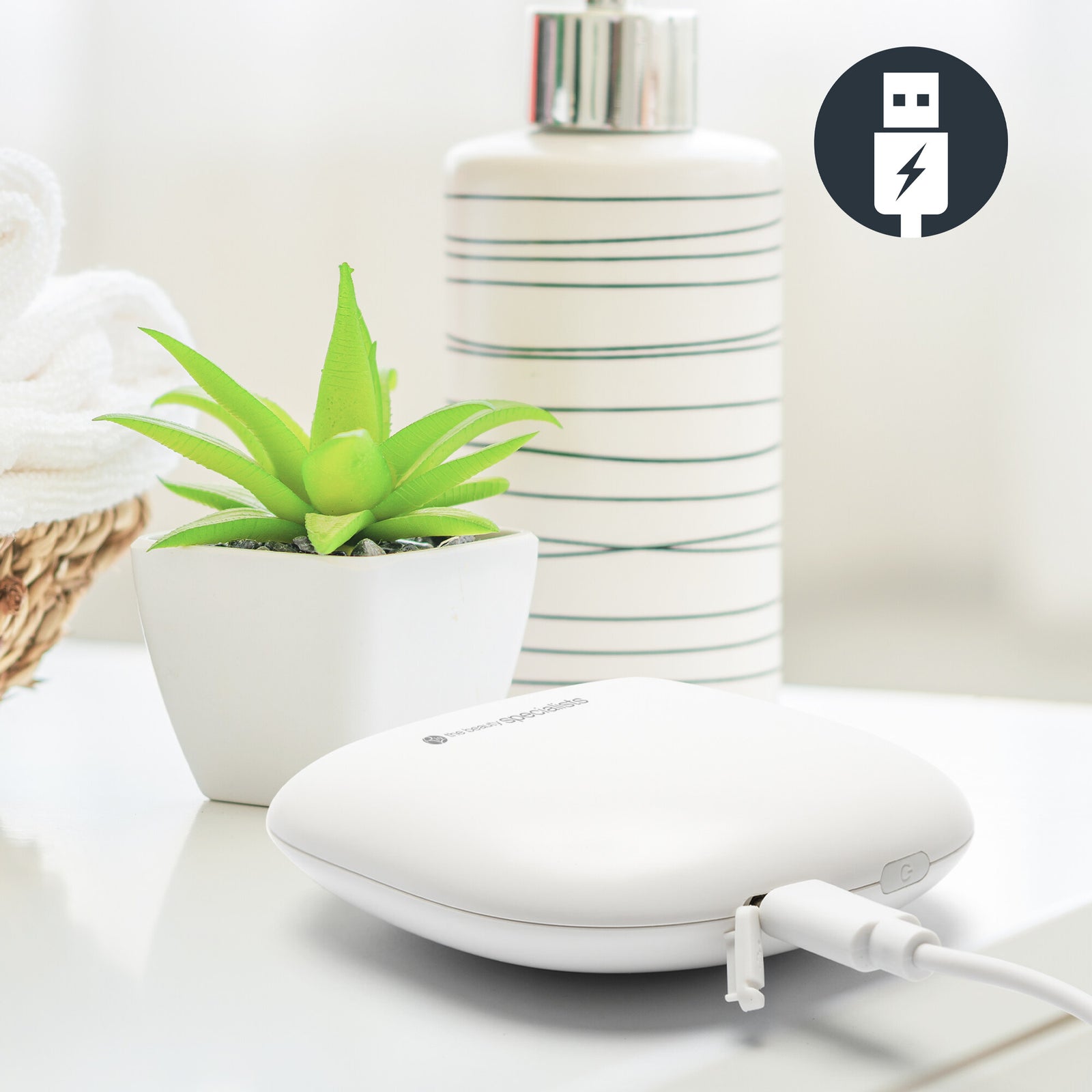 The Rio Face Lift & Tone Facial Toner, a compact white device, charges via USB on a countertop beside a potted succulent and striped bottle. A USB charging icon appears in the corner. Ideal for skin toning and anti-aging routines.