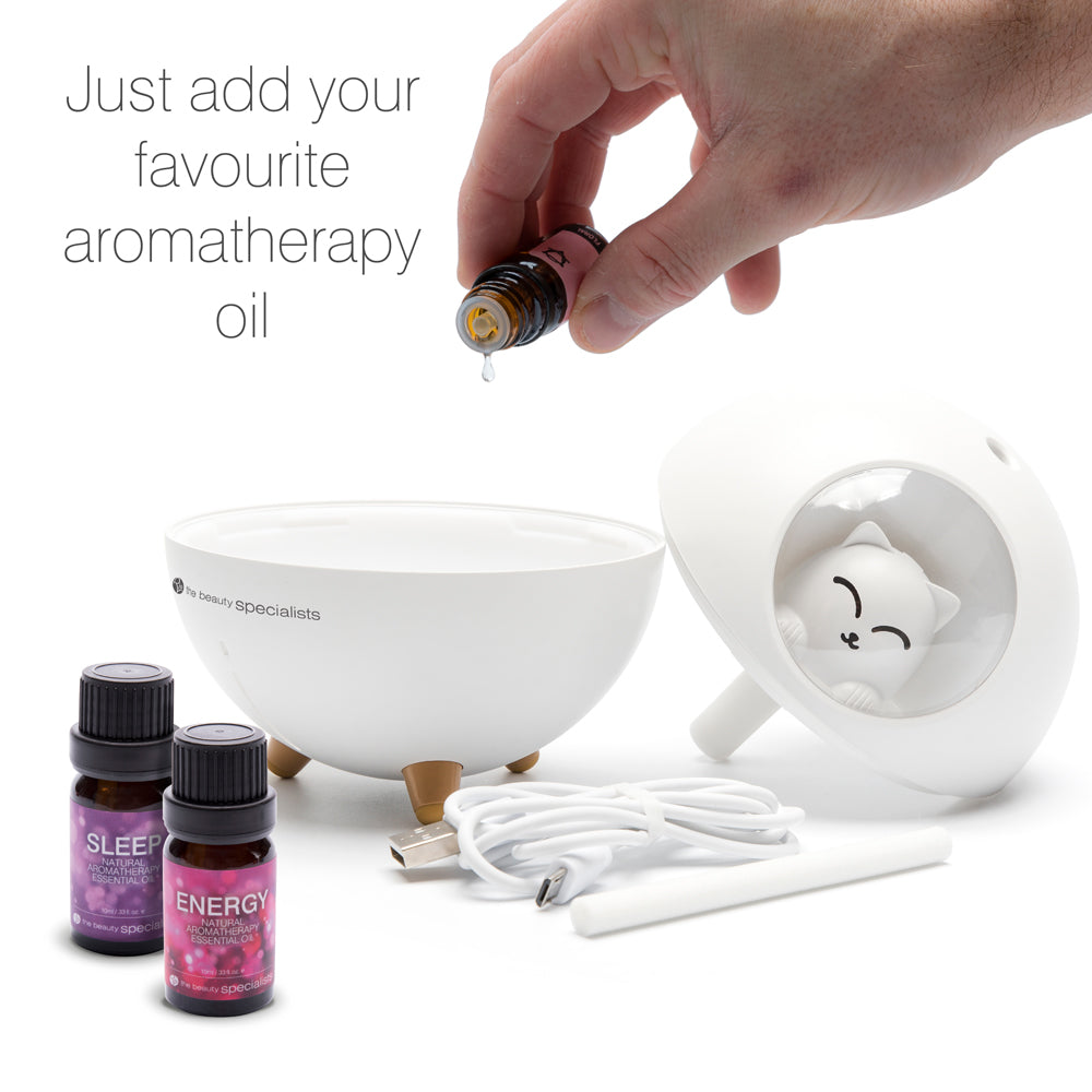 A person adds aromatherapy oil to the Rio MIA Cat Children’s Essential Oil Diffuser, Humidifier & Night Light. Bottles labeled SLEEP and ENERGY, a USB cable, and diffuser parts are nearby. Text: Add your favourite oil for night-light bliss.