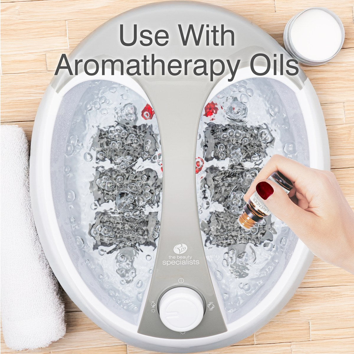 A hand adds aromatherapy oil to a bubbling Rio Deluxe Foot Spa Bath and Massager filled with water. A towel and jar rest on a bamboo mat. Text on the image reads: Use With Aromatherapy Oils.