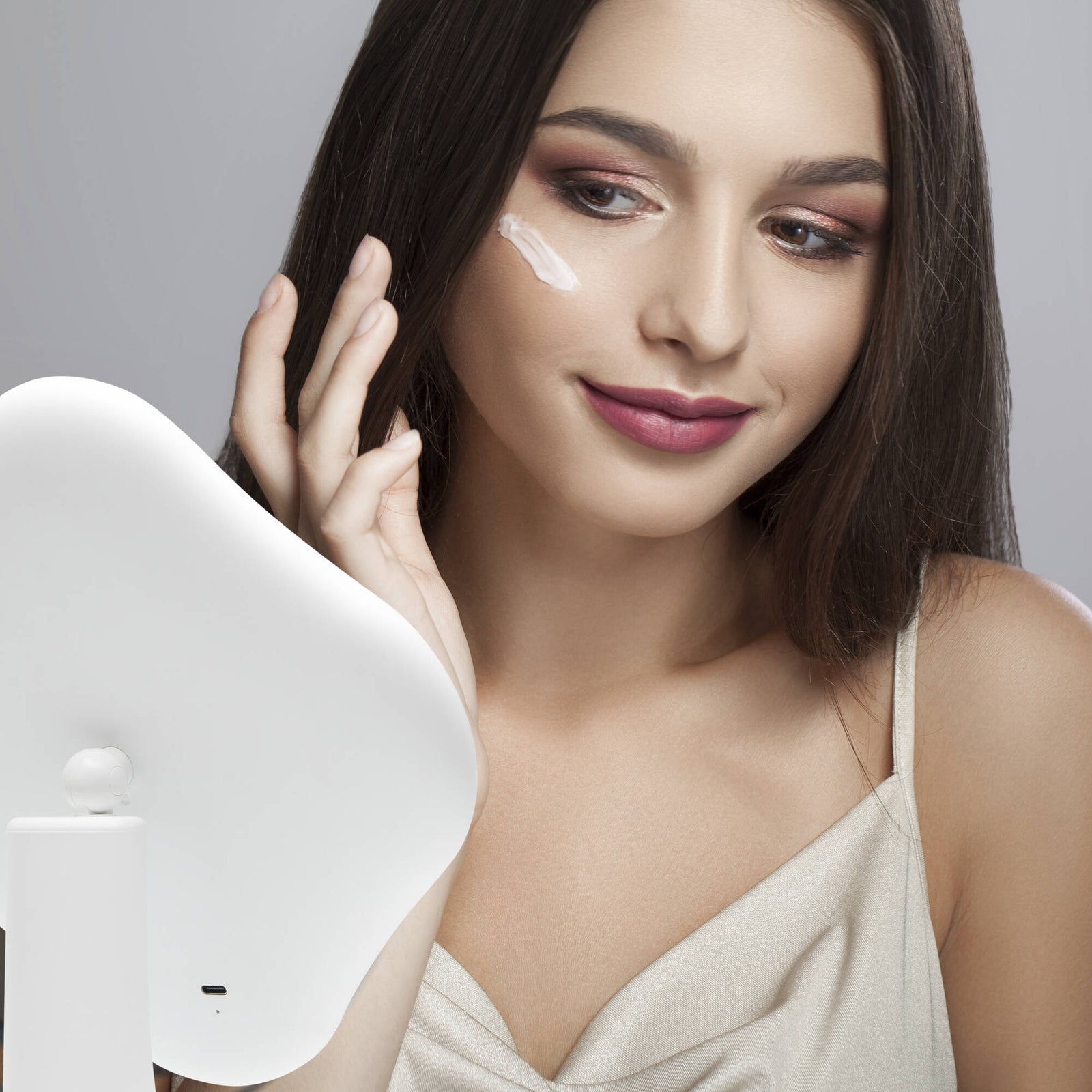 A young woman with long dark hair, wearing a light sleeveless top, smiles gently as she applies cream to her cheek while looking into the Rio Illuminated Full Size Beauty Mirror.