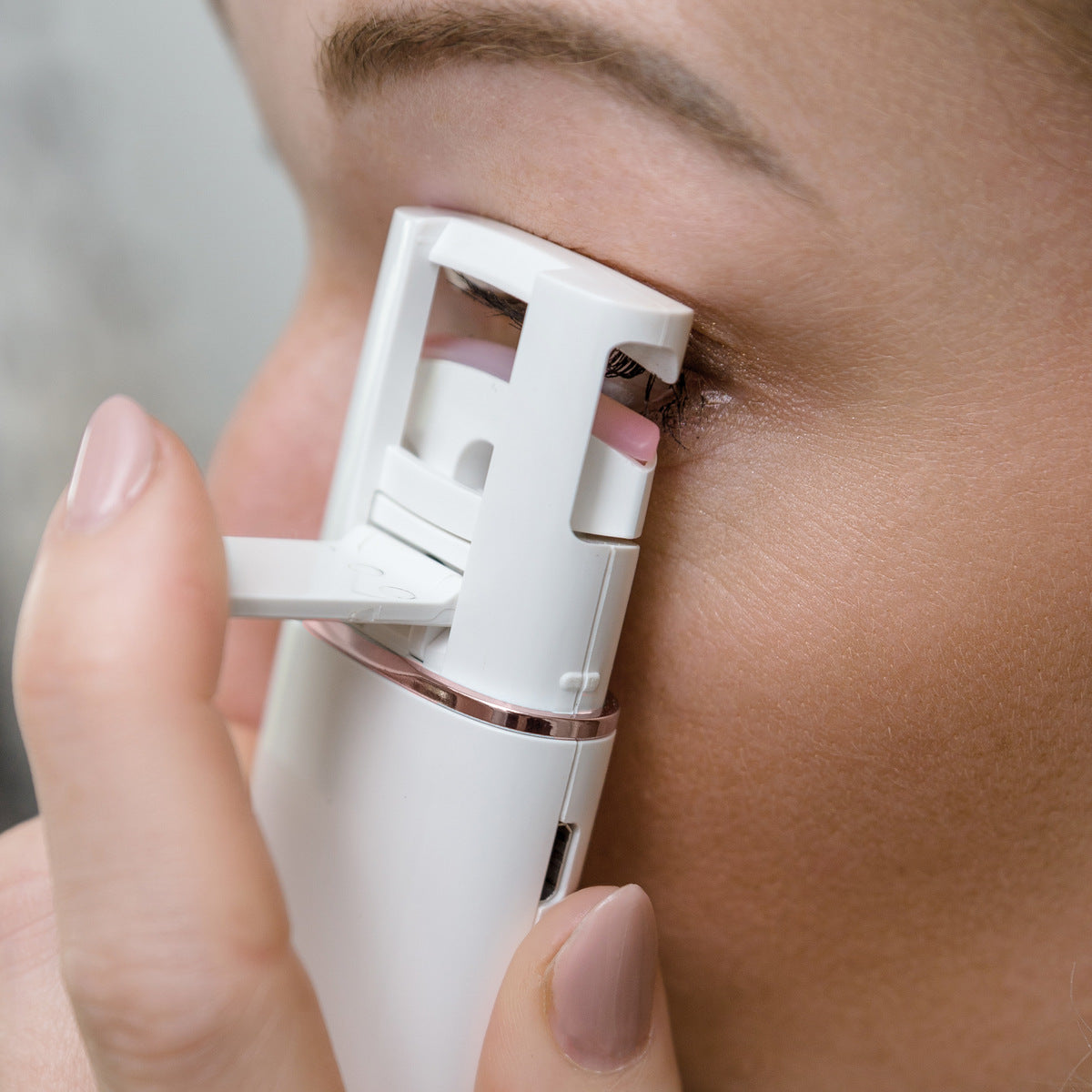 A close-up shows a person using the Rio WOW Lash Heated Eyelash Curler on their upper lashes, focusing on the eye and curler, with neatly manicured fingers holding the device for beautifully curled lashes.