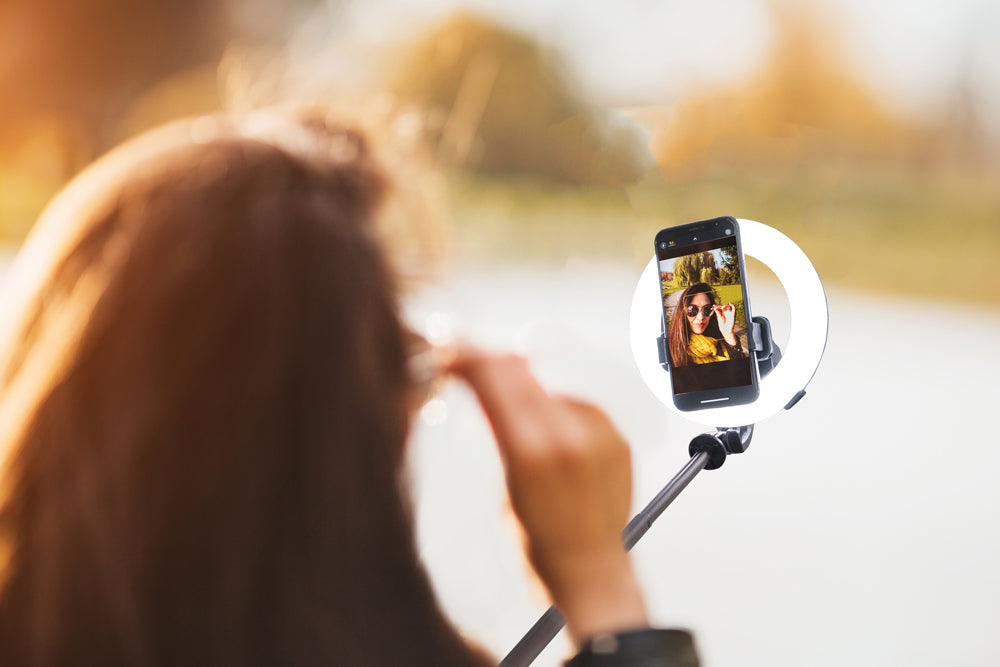 A woman poses for a selfie outdoors with the Rio GlamourGlow Pro Beauty & Vlogging Ring Light. Her smartphone, mounted on the ring light, displays her smiling against a backdrop of blurred greenery—ideal for vlogging and flawless photos.