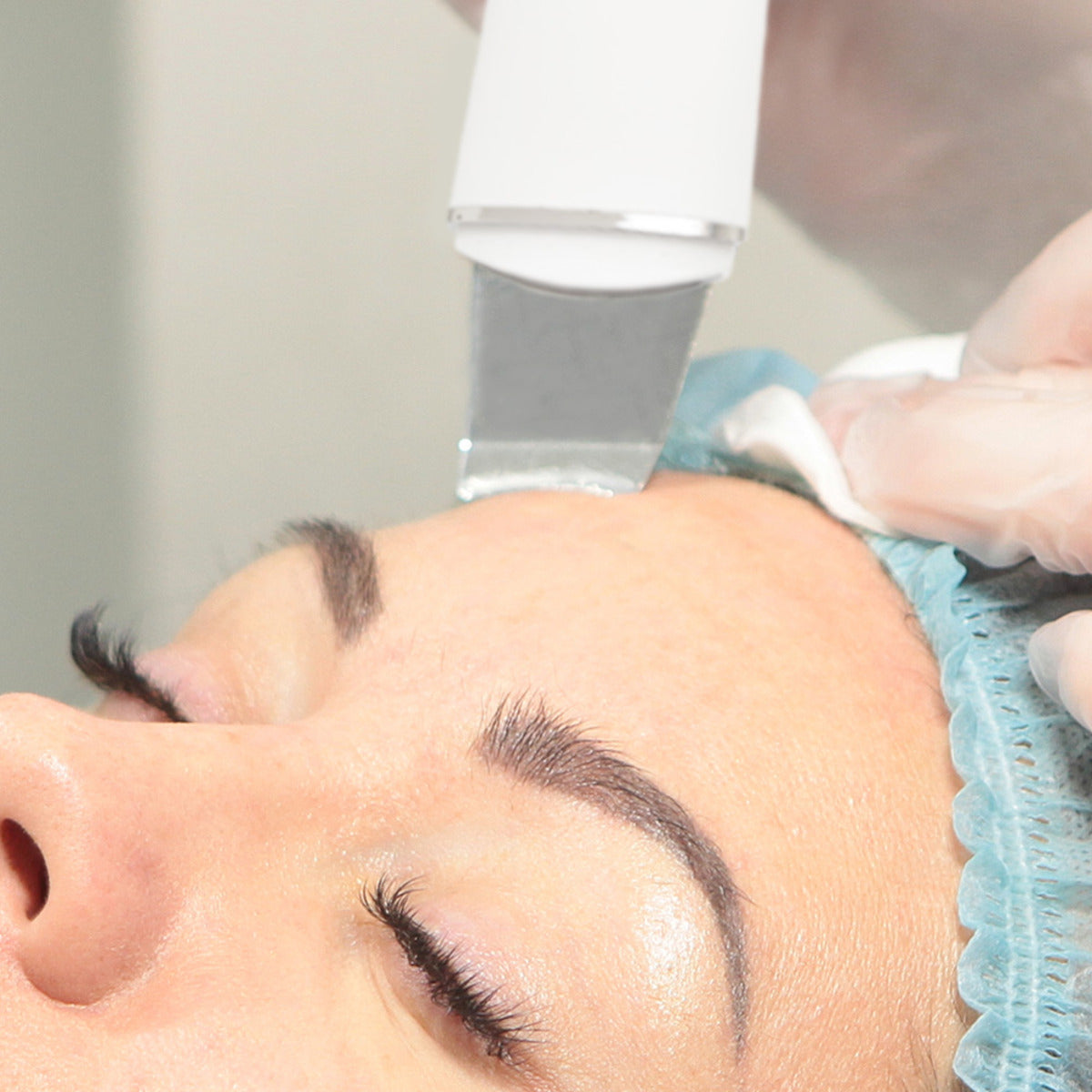 A person with closed eyes and a blue medical cap receives a skin treatment using the Rio Ultrasonic Skin Cleanser, shown in close-up as the device glides over their forehead.