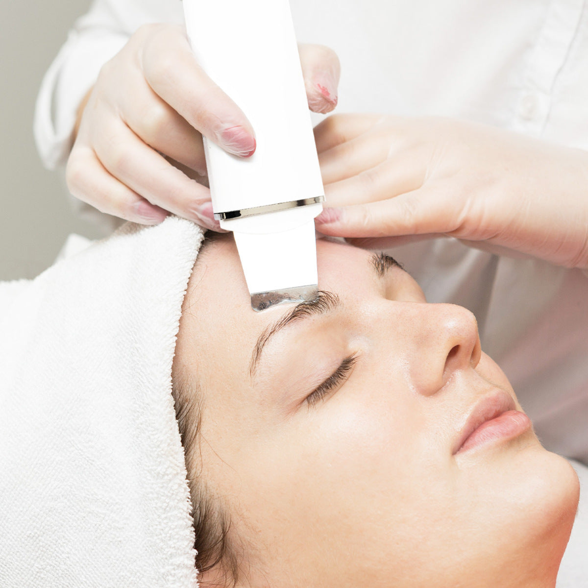 A person with a towel around their head receives a facial treatment as a gloved professional uses the Rio Ultrasonic Skin Cleanser on their forehead.