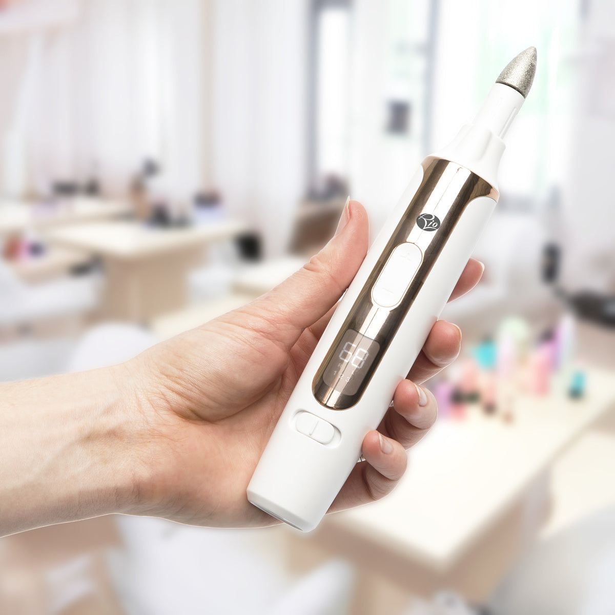 A hand holds the Rio Precision Pro Electric Nail File, a white and gold device with a digital display, highlighting precise professional nail care in a bright, blurred salon background.