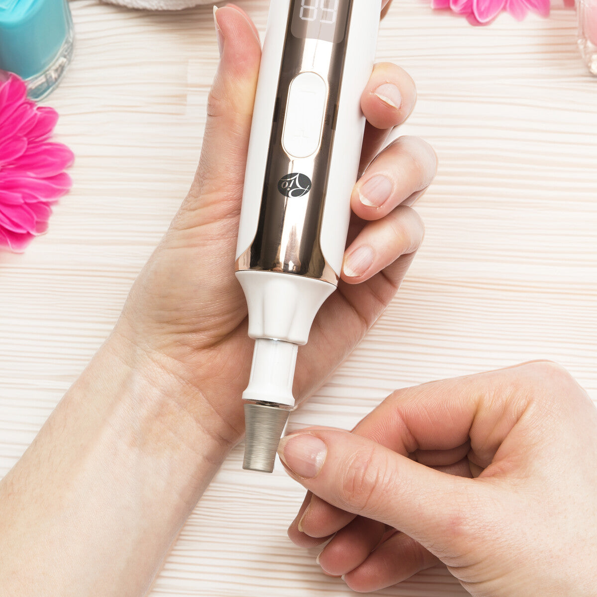 A person attaches a nozzle to the Rio Precision Pro Electric Nail File with precision, holding it in one hand and fitting the attachment with the other on a light wooden surface, surrounded by pink flowers and nail care items.
