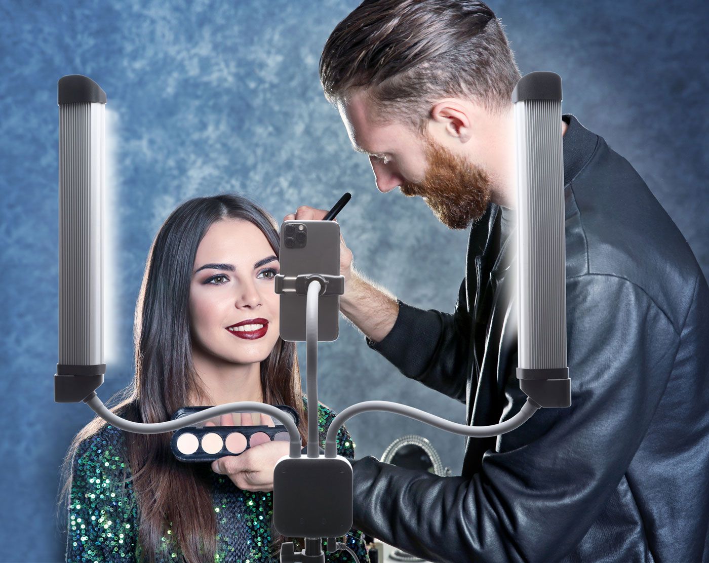 A makeup artist applies eyeshadow to a smiling woman as Rio Professional Twin LED Strip Lights illuminate the scene, while ring lights and a phone on flexible stands capture content against a textured blue background.