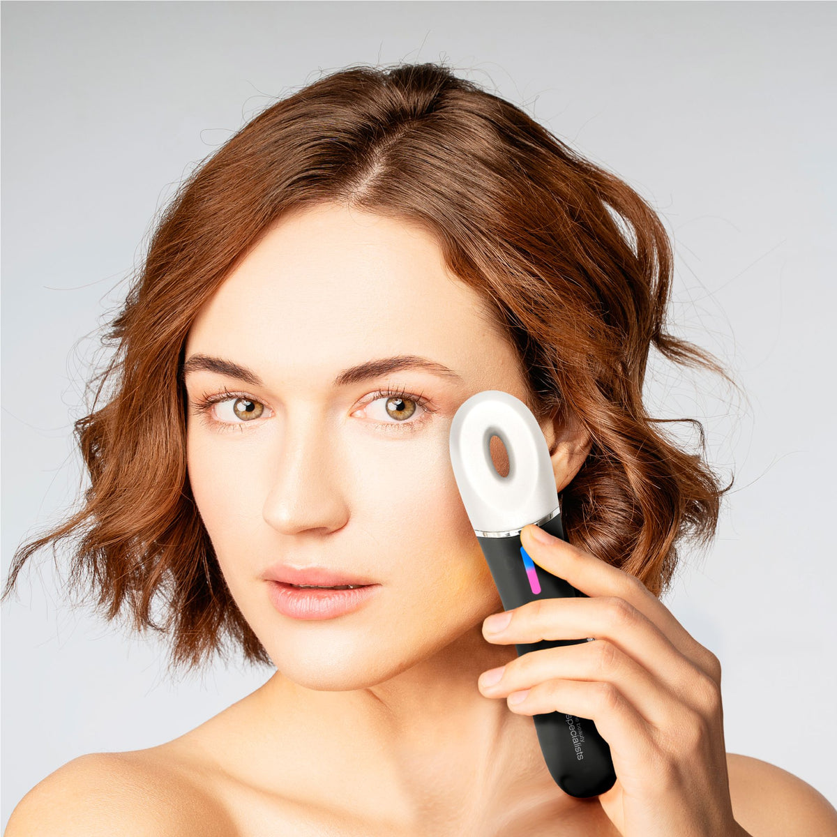 A woman with short brown hair uses the Rio Revive & Refresh handheld electronic skincare device on her cheek, looking at the camera with a neutral expression against a plain, light background.