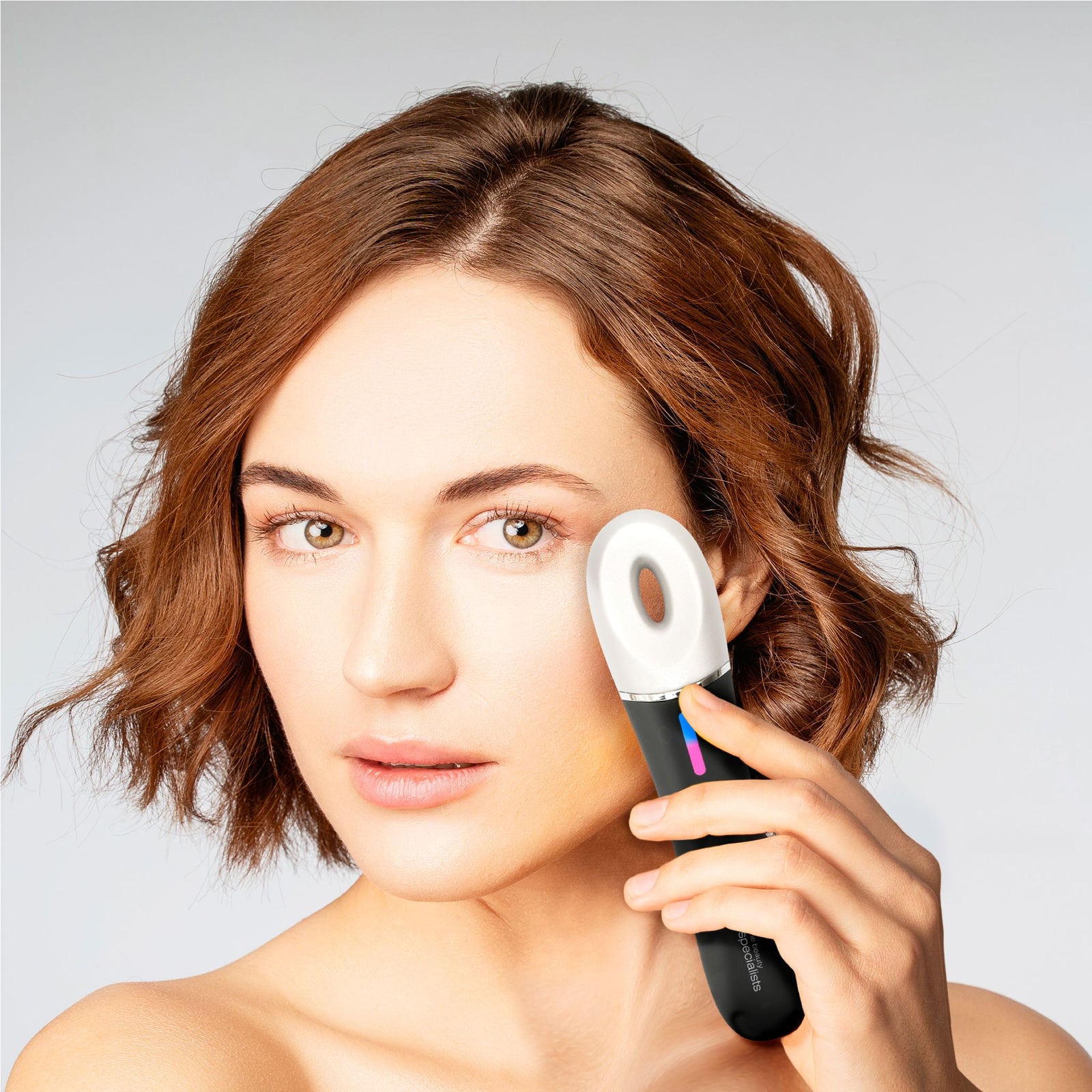 A woman with short brown hair uses the Rio Revive & Refresh handheld electronic skincare device on her cheek, looking at the camera with a neutral expression against a plain, light background.