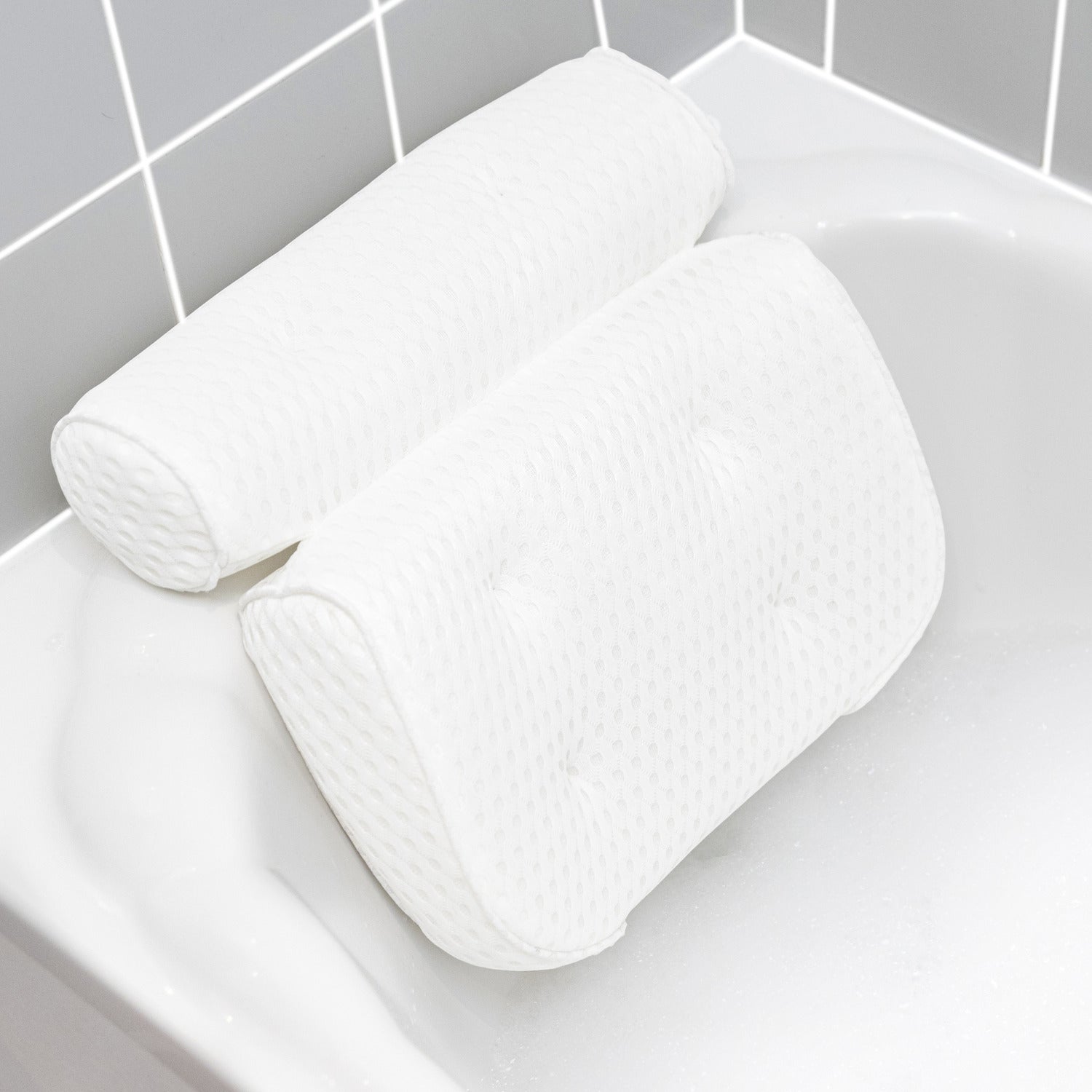 The Rio Luxury Bath Spa Pillow, featuring a white textured design with two plush cushions, rests on the edge of a white bathtub beside gray and white tiled walls.
