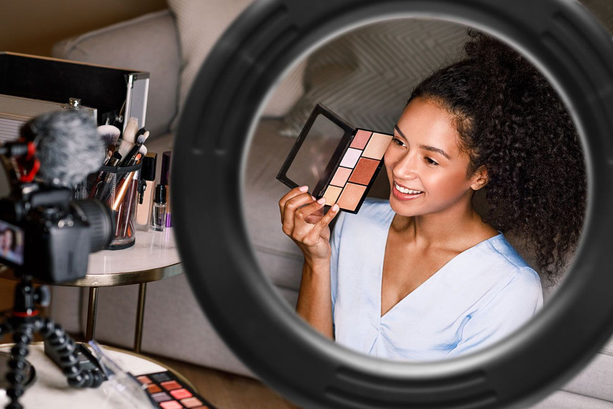 A woman smiles and films a beauty tutorial at home, demonstrating flawless makeup while using the Rio Makeup Perfector LED Ring Light, surrounded by her makeup products.