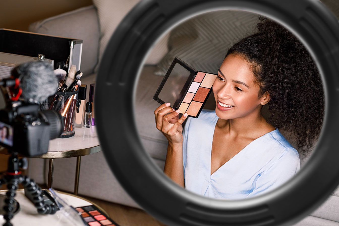 A woman smiles and films a beauty tutorial at home, demonstrating flawless makeup while using the Rio Makeup Perfector LED Ring Light, surrounded by her makeup products.