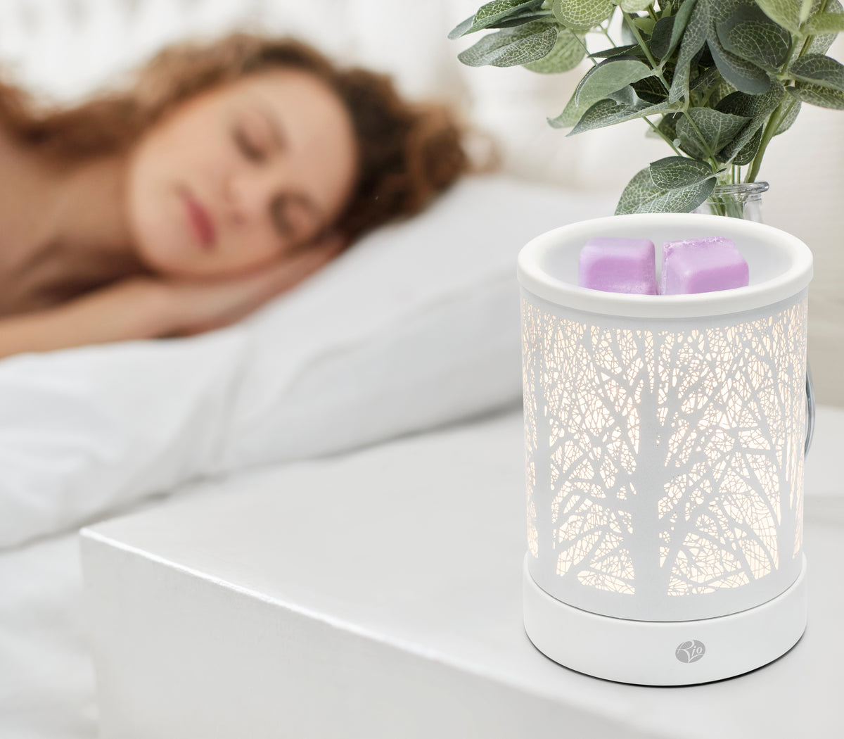 A woman sleeps on a white bed beside a nightstand with the Rio FORA Wax Melt Burner & Aroma Diffuser Lamp glowing softly, holding two purple wax melts and a green potted plant, letting her relax in style.