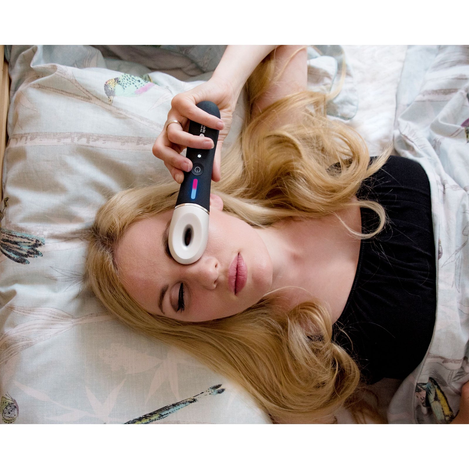 A woman with long blonde hair relaxes on bird-patterned bedding, eyes closed, using the Rio Revive & Refresh handheld skincare device on her forehead. She wears a black top.